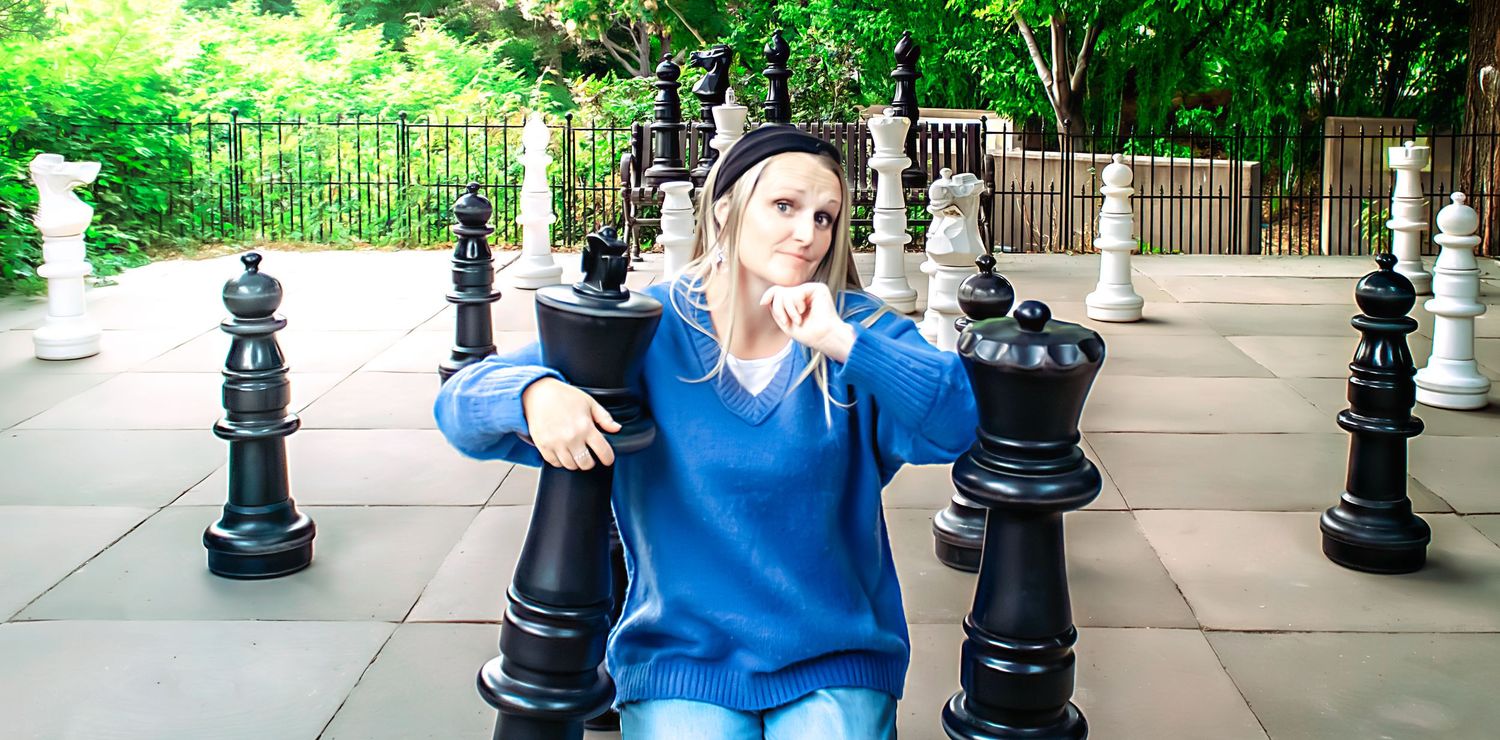 A person in blue plays with oversized outdoor chess pieces in a garden setting.