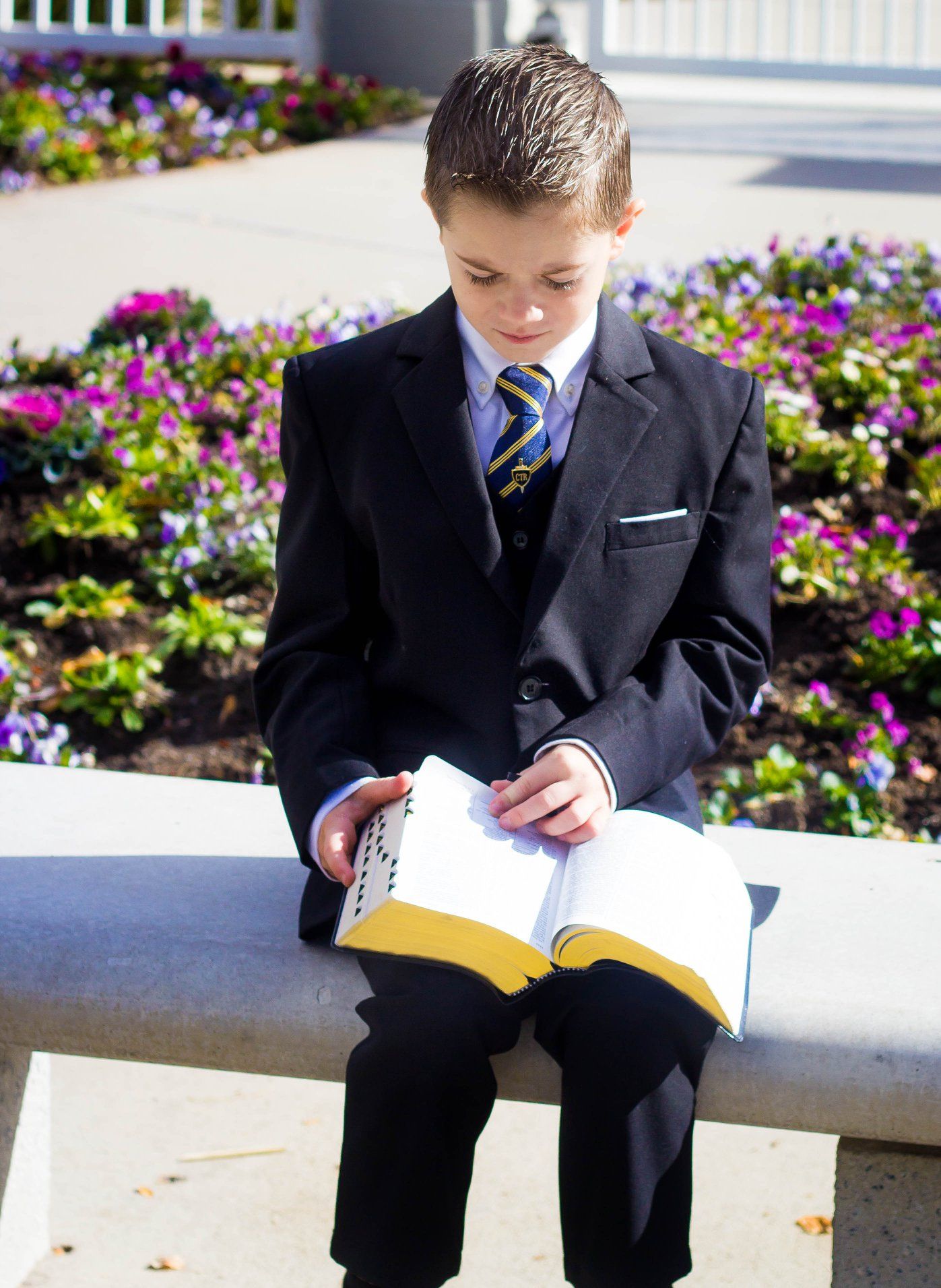 Child in dark suit reading book while sitting on outdoor ledge with flowers.