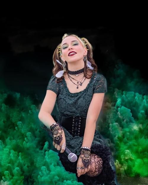 Performer in black lace dress with microphone against dramatic green smoke background.