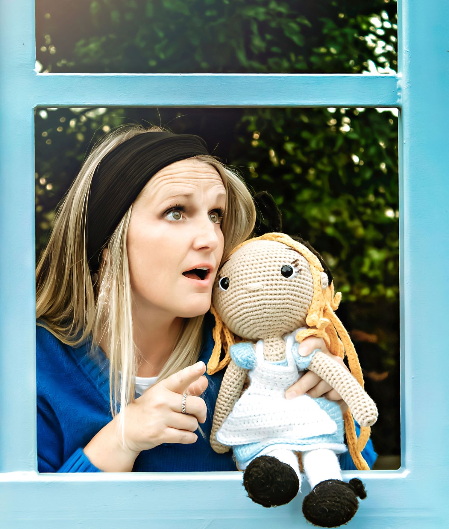 A person poses playfully with a crocheted doll through a light blue window frame.