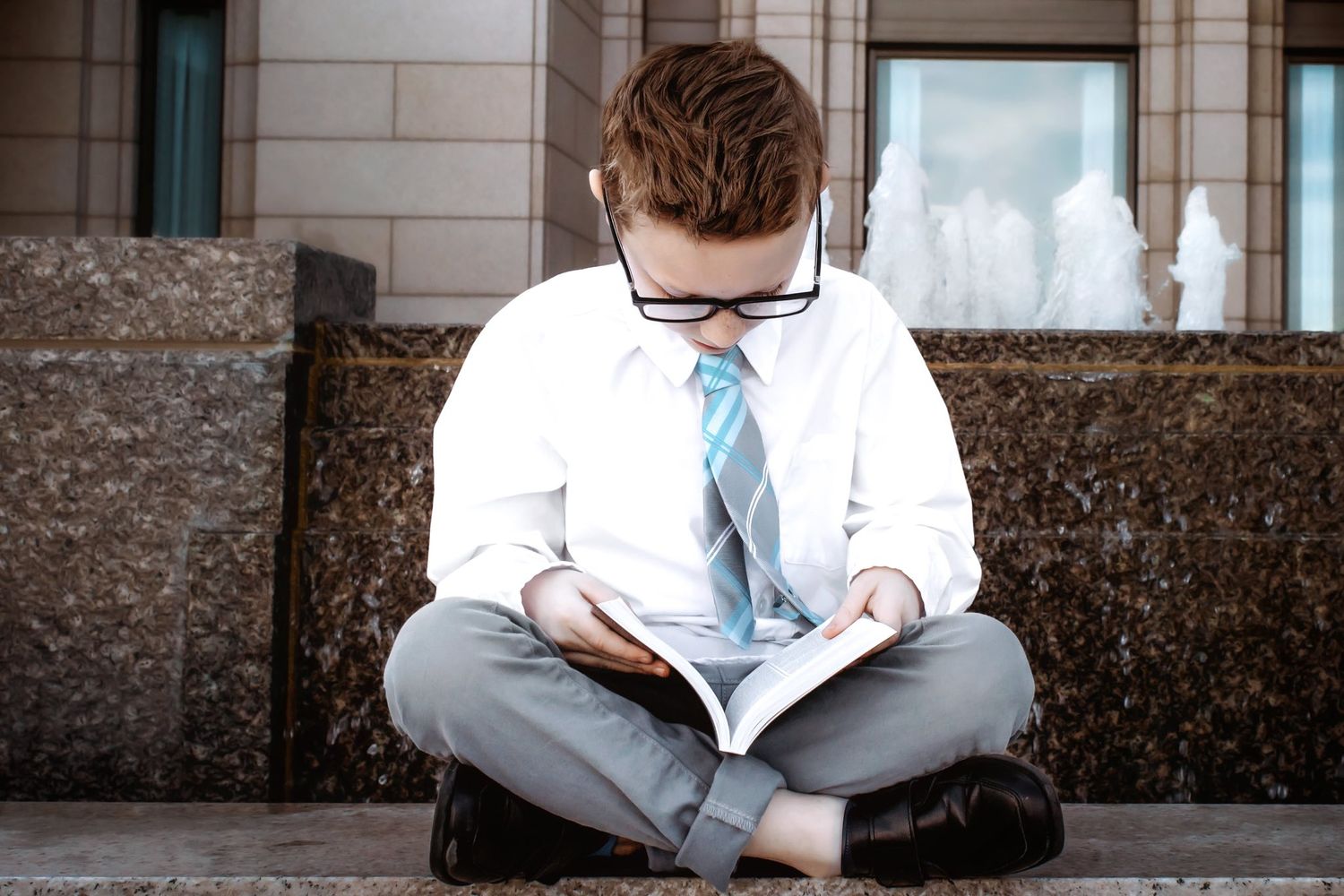 Person in formal attire sitting on stone ledge reading a book.