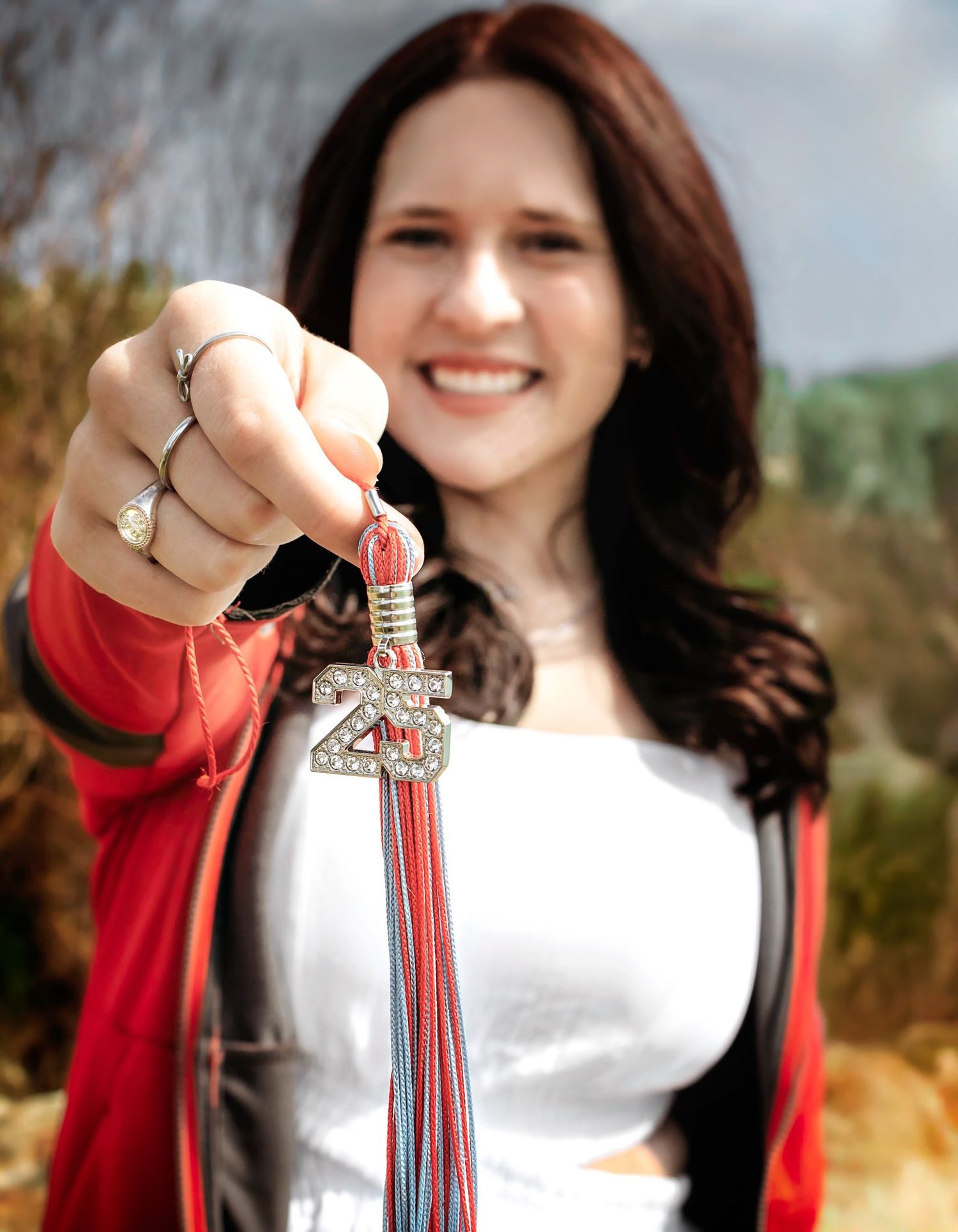 Person in white top and red cardigan holding graduation tassel outdoors.