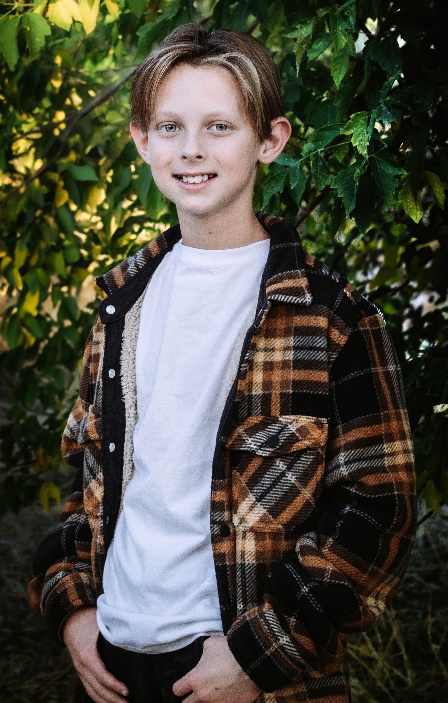 A child in plaid flannel and white shirt poses confidently against green leaves.