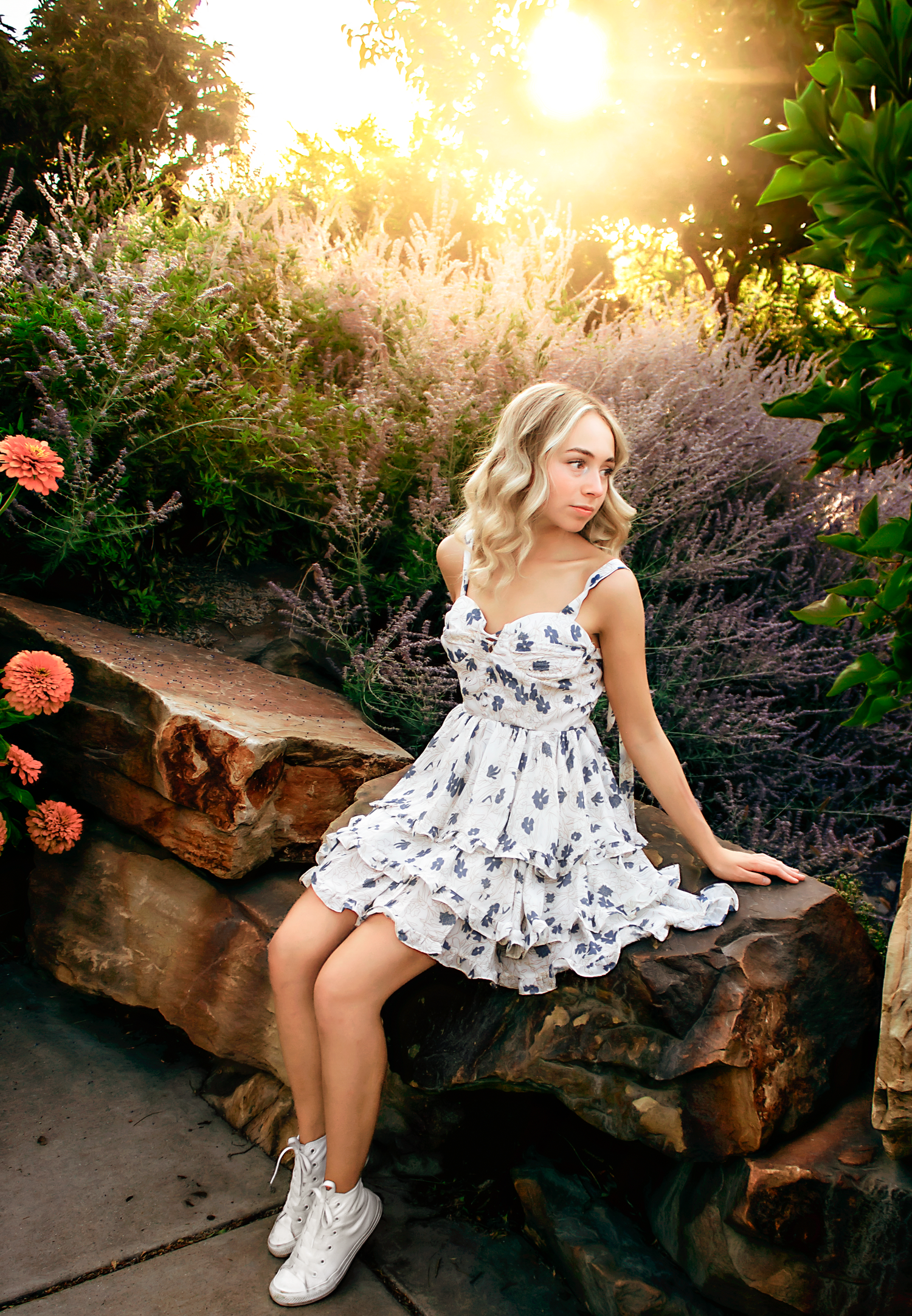 Person in floral dress sitting on stone wall with garden backdrop during golden hour.