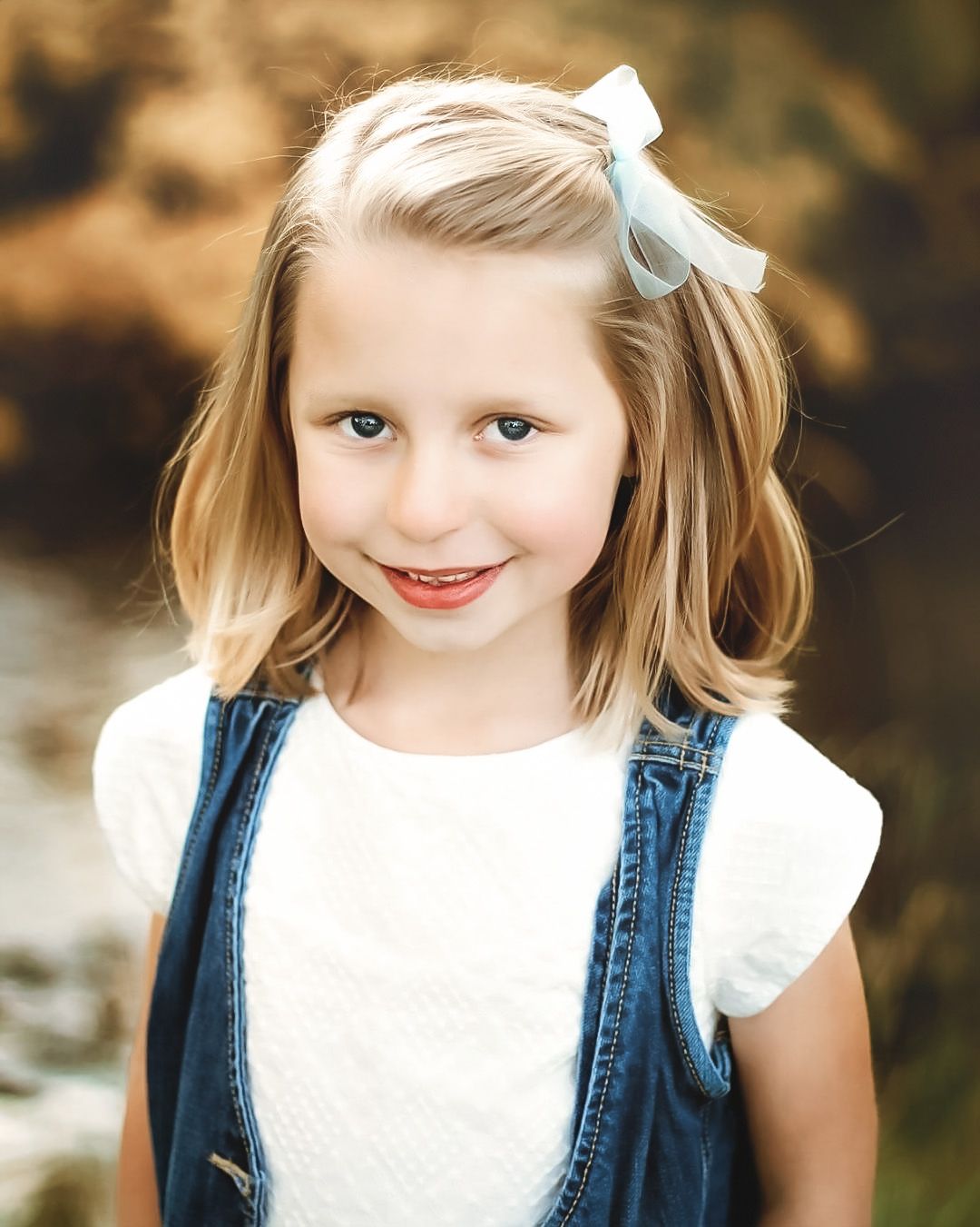 Child wearing denim and white top poses outdoors in autumn setting.