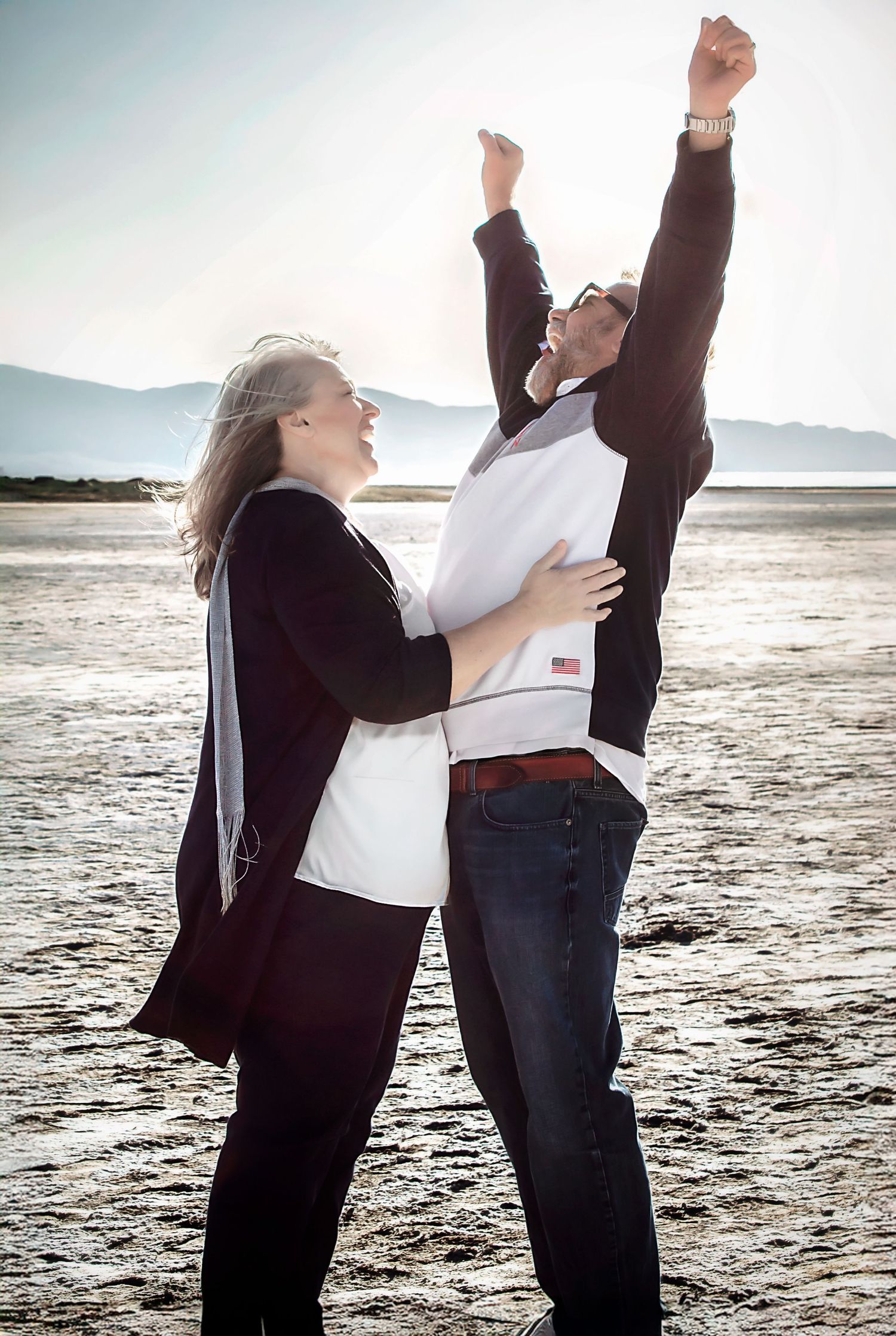 Couple dances joyfully together on sandy beach with mountains visible in distance.