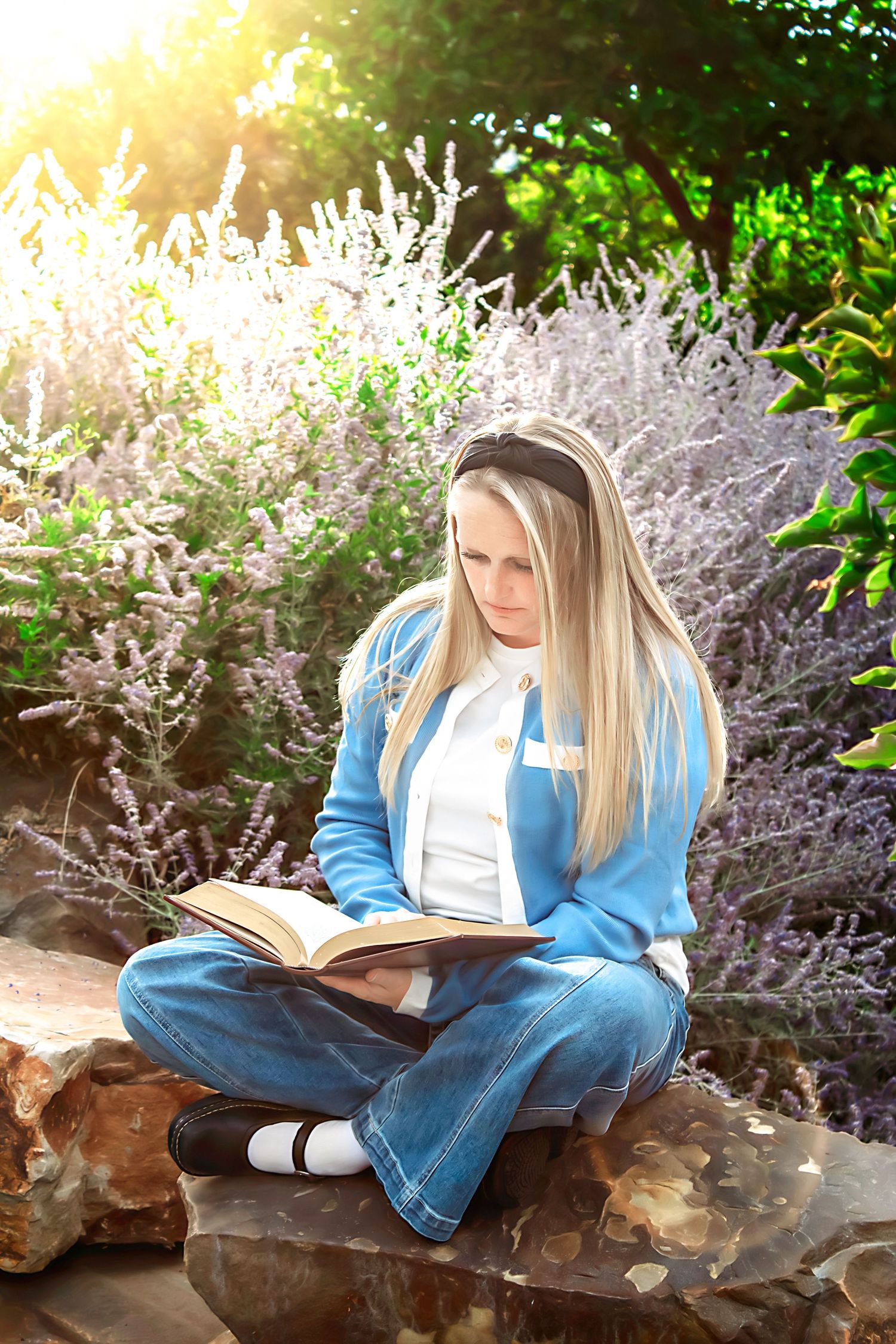 Person in denim reading a book while resting on natural rock formation amid flowering plants.