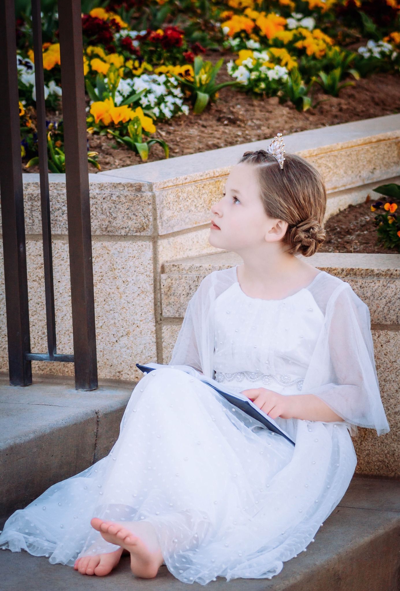 Person in white dress sitting on concrete steps near colorful flower garden.