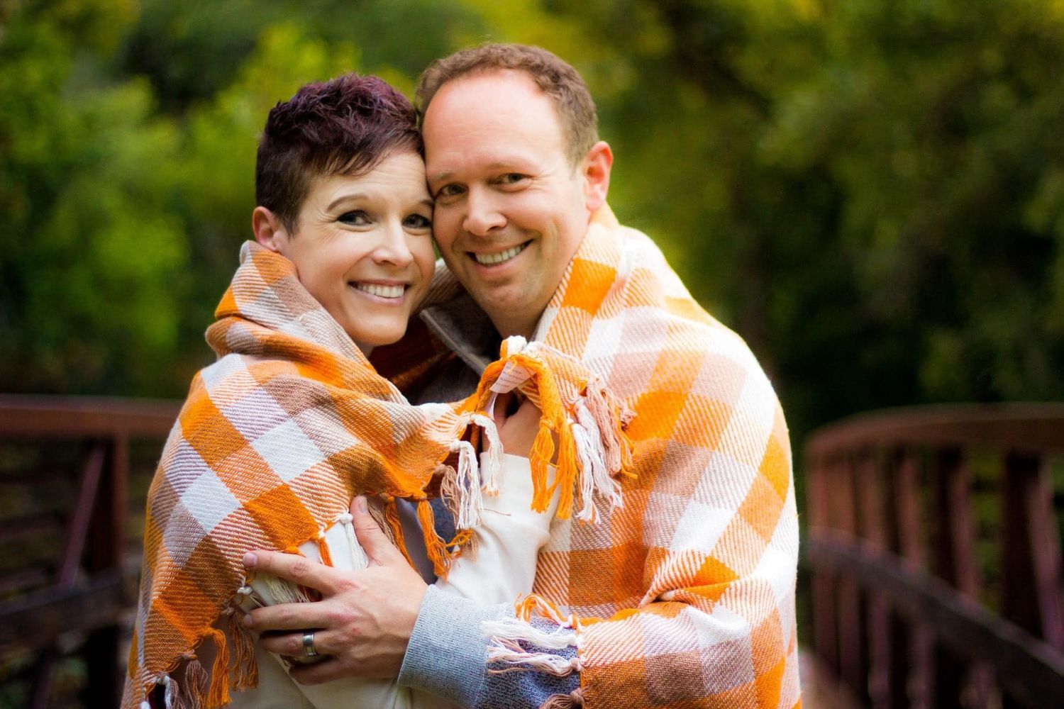 Couple wrapped in orange blanket sharing intimate moment on wooden bridge.