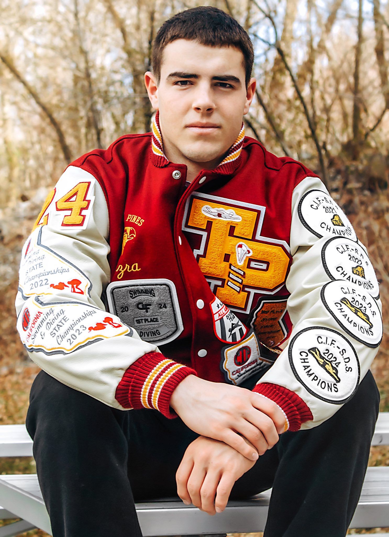 A student wearing a red and white varsity letterman jacket with patches and emblems sitting outdoors.