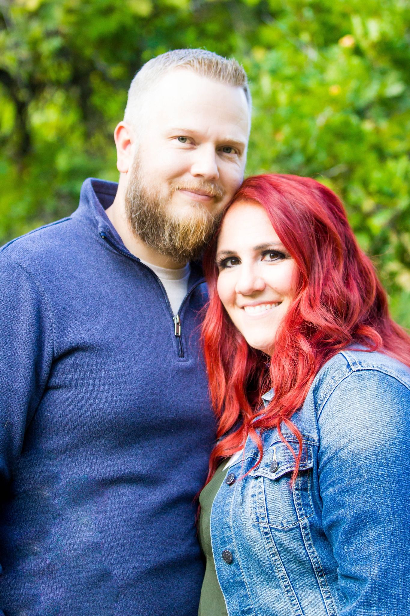 Couple in casual wear smiling together against green foliage background.