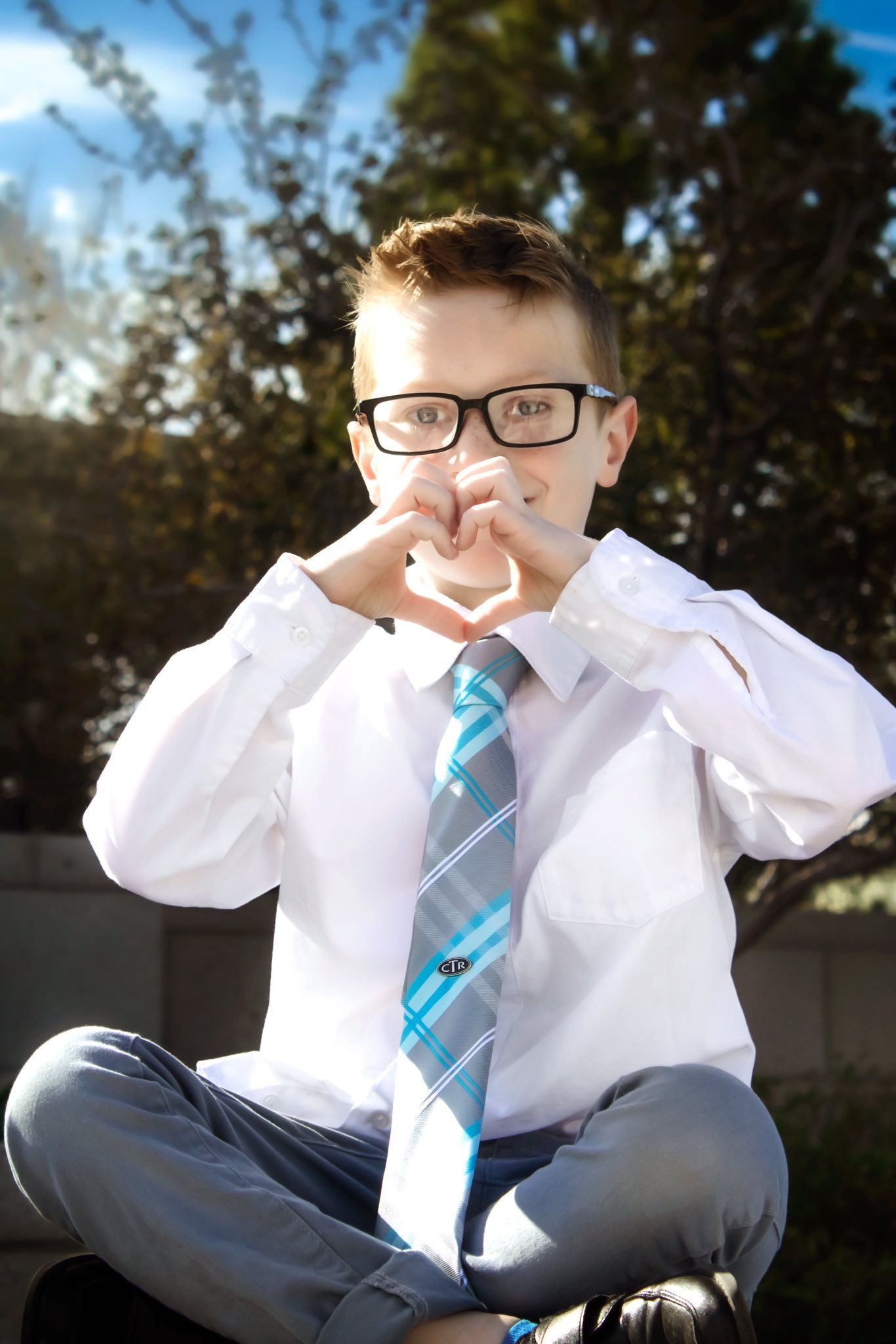 Child in formal attire with glasses making playful expressions outdoors.