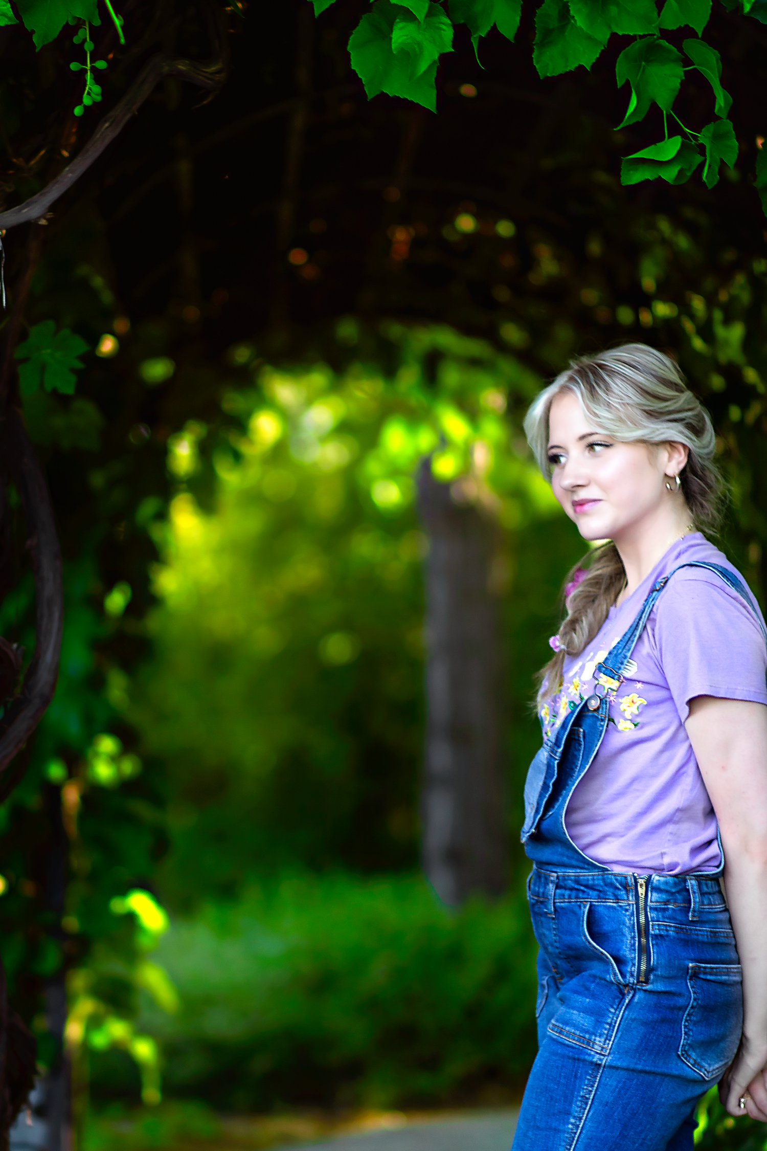 A casual outdoor photoshoot featuring denim overalls and a purple t-shirt against a lush green background.