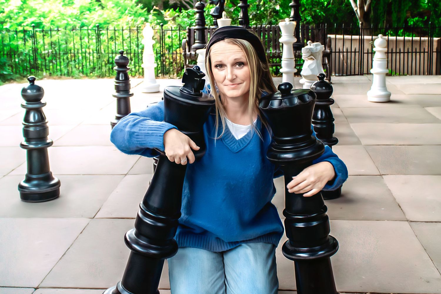 Person in casual attire sits between large decorative chess pieces on stone path.