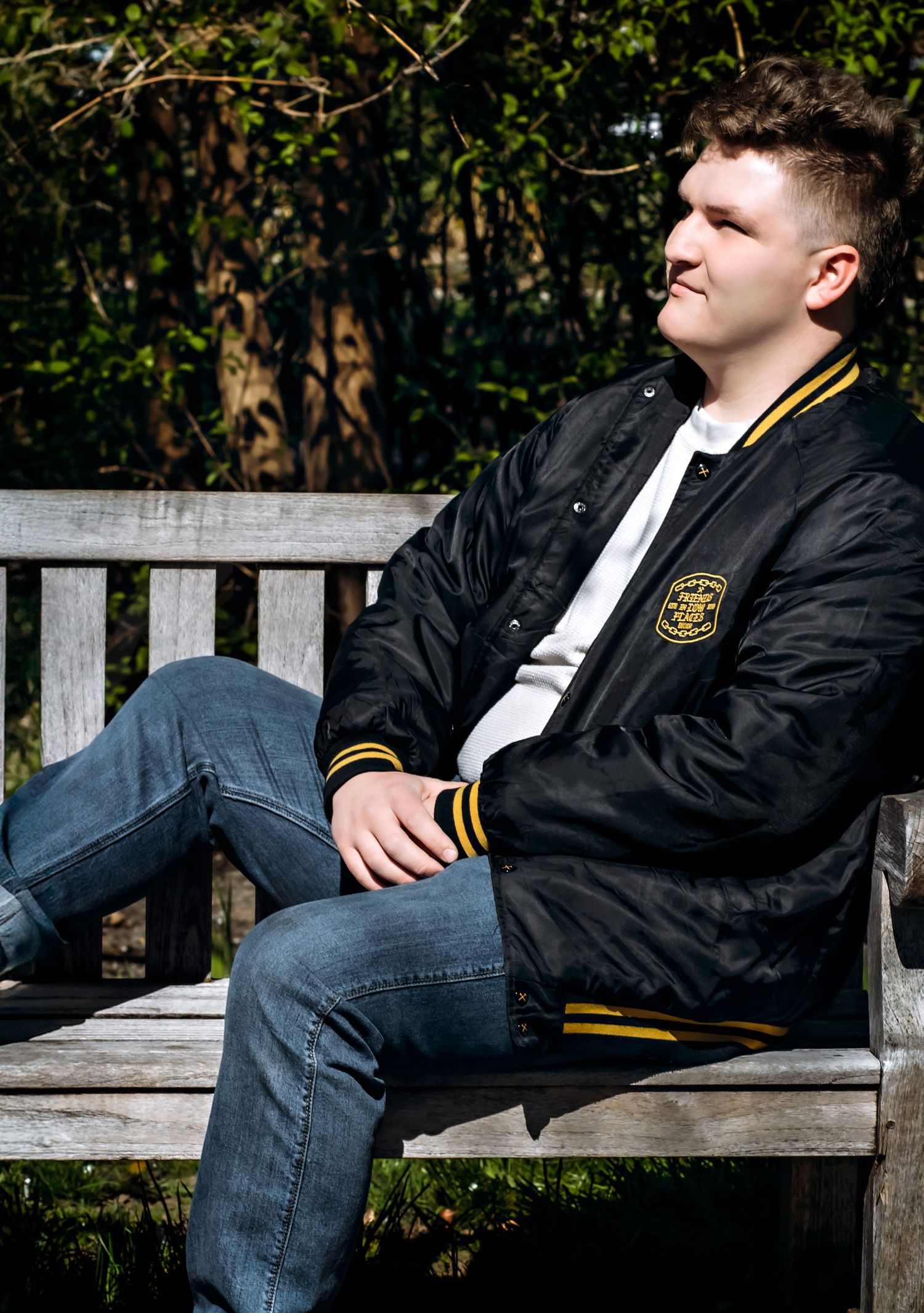 Person relaxing on wooden bench wearing black and gold varsity jacket in garden setting.