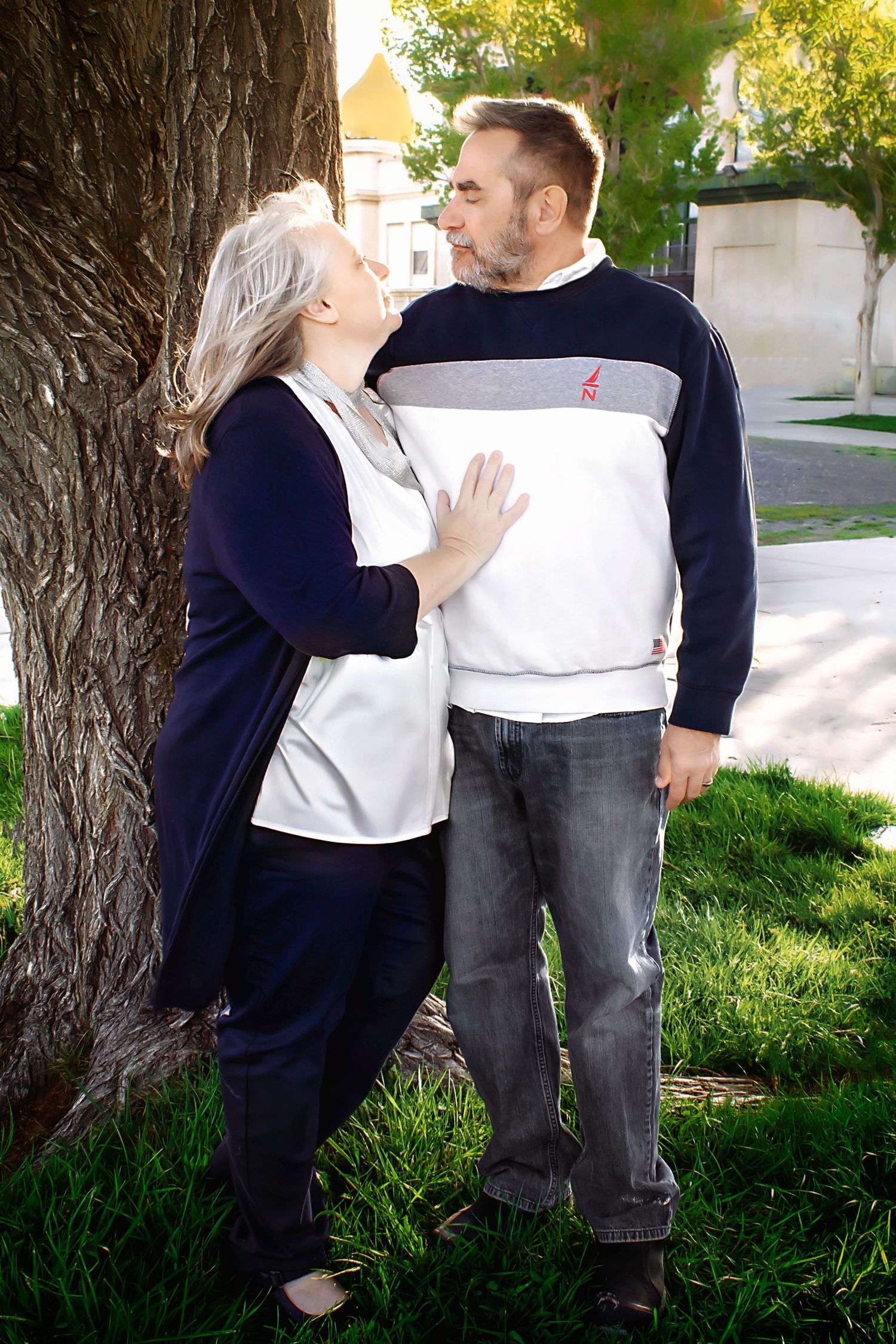Romantic couple poses together near large tree trunk in sunny outdoor setting.