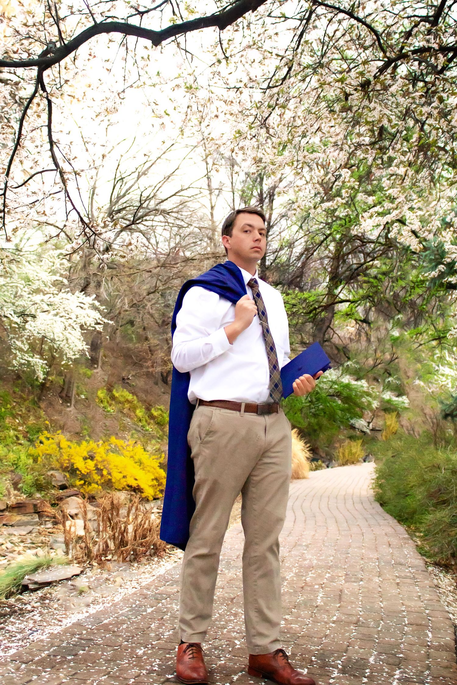 Graduate in formal attire poses on a brick path surrounded by blooming spring trees.