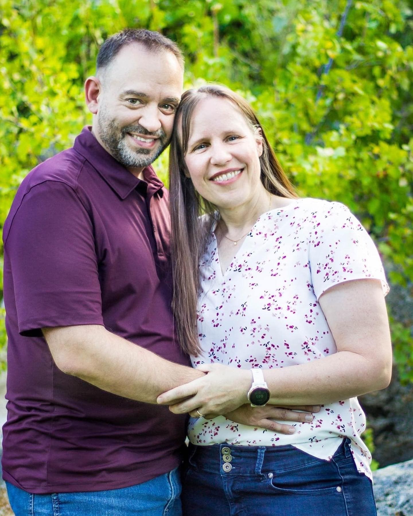 Couple in casual attire embracing outdoors with green summer foliage background.