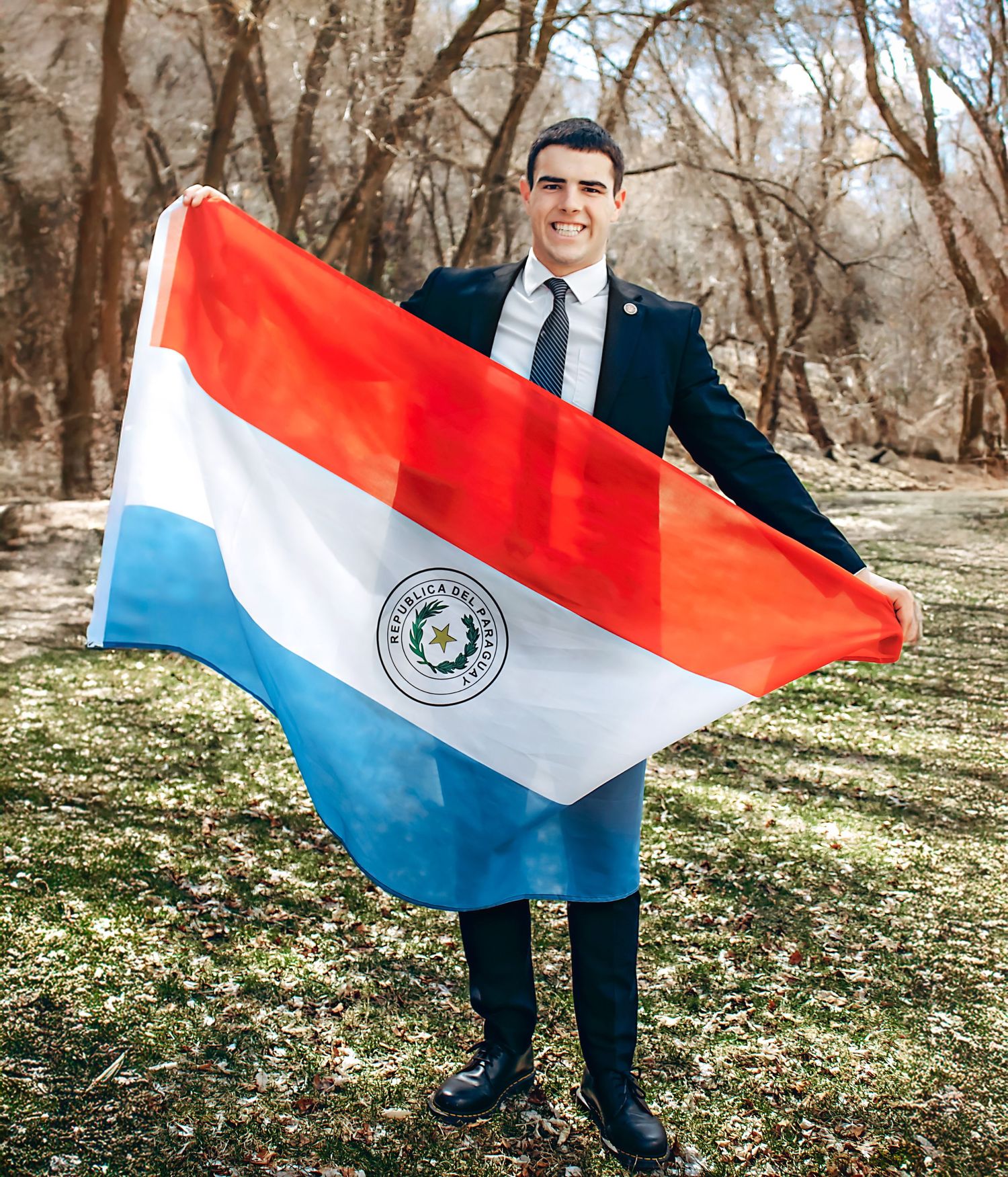 Person holding a red, white and blue flag outdoors in a wooded area.