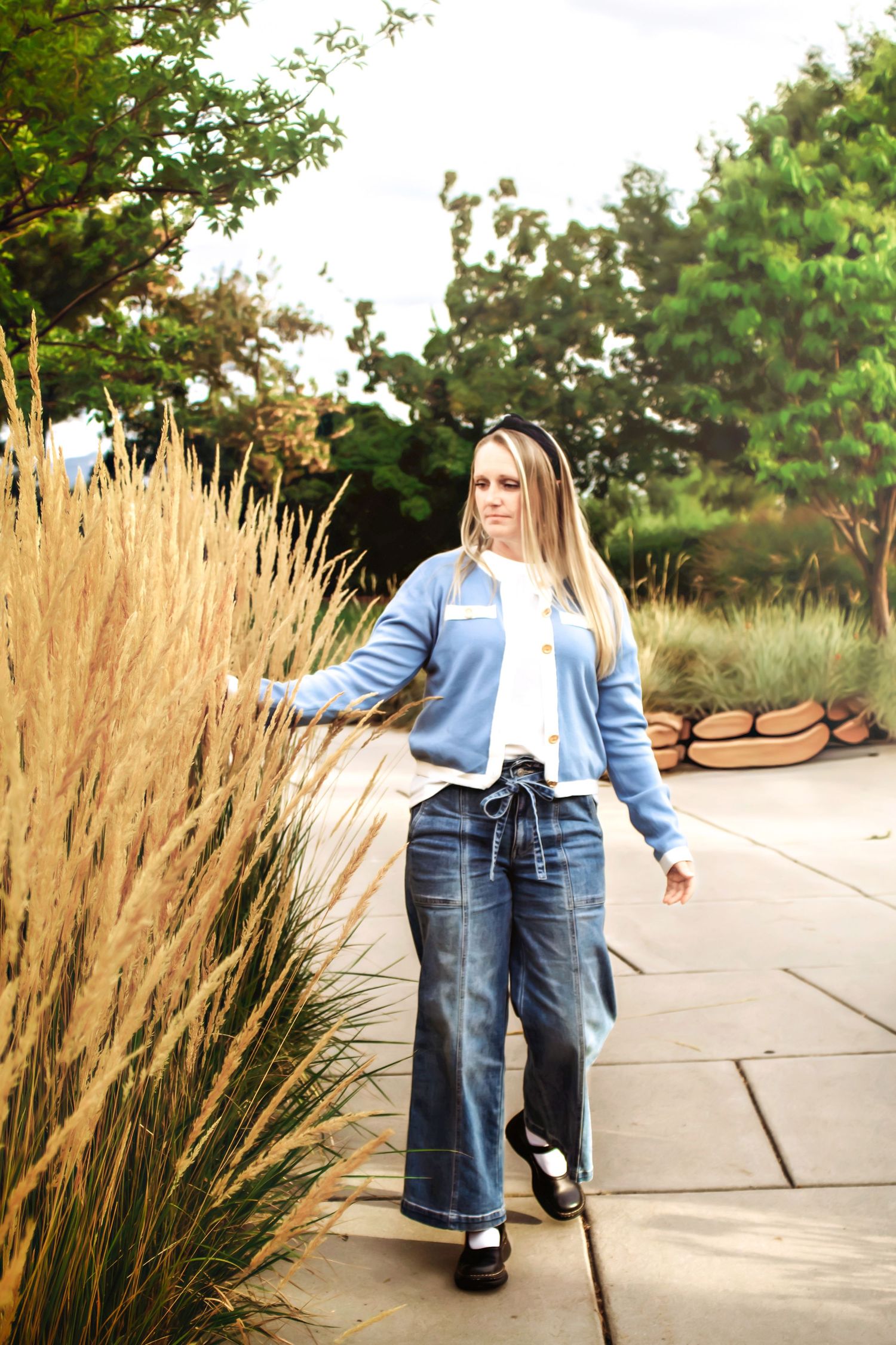 Person wearing light blue jacket and jeans poses against ornamental grasses in outdoor garden setting.