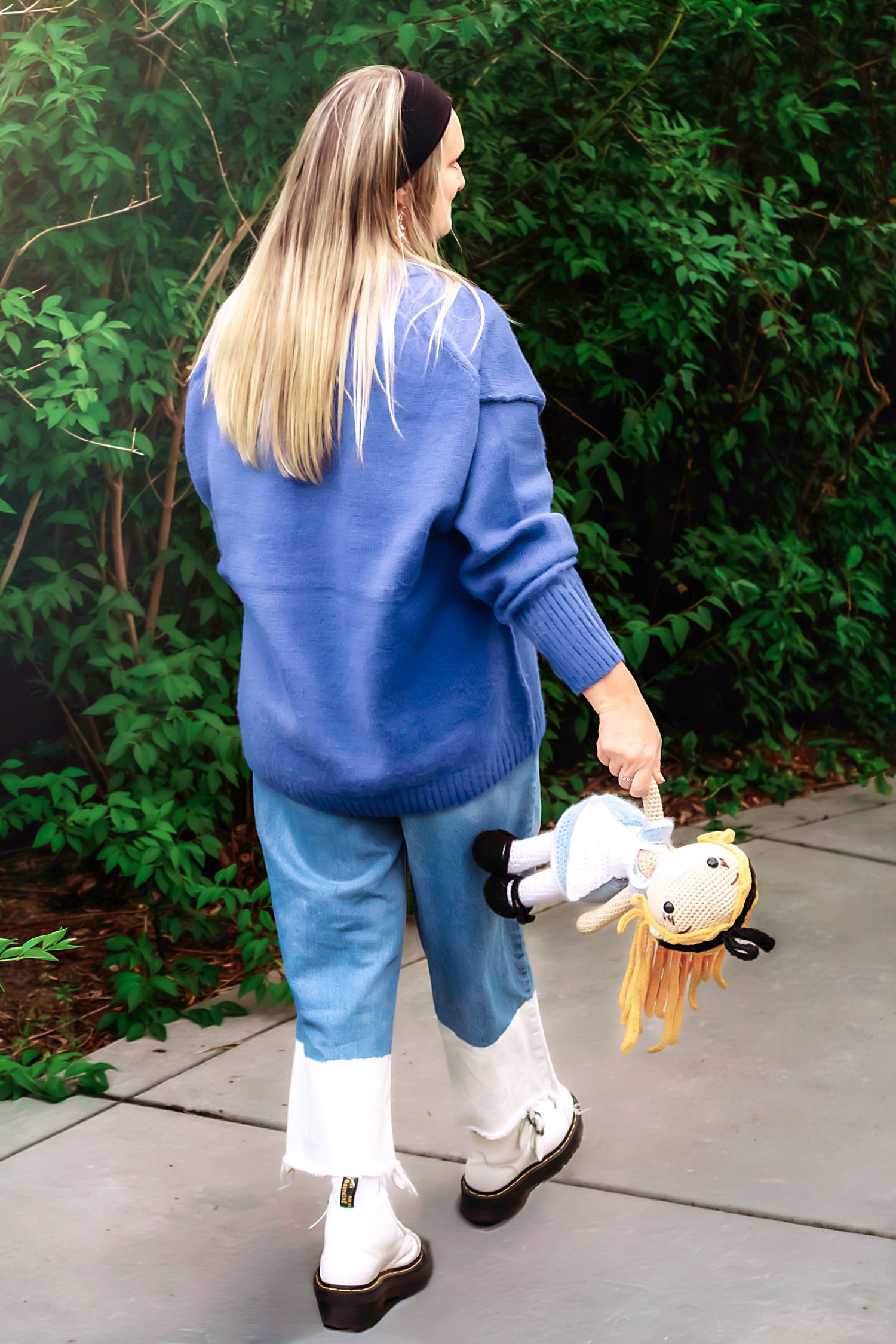 A person in a blue sweater walks along a concrete path carrying a small plush toy.