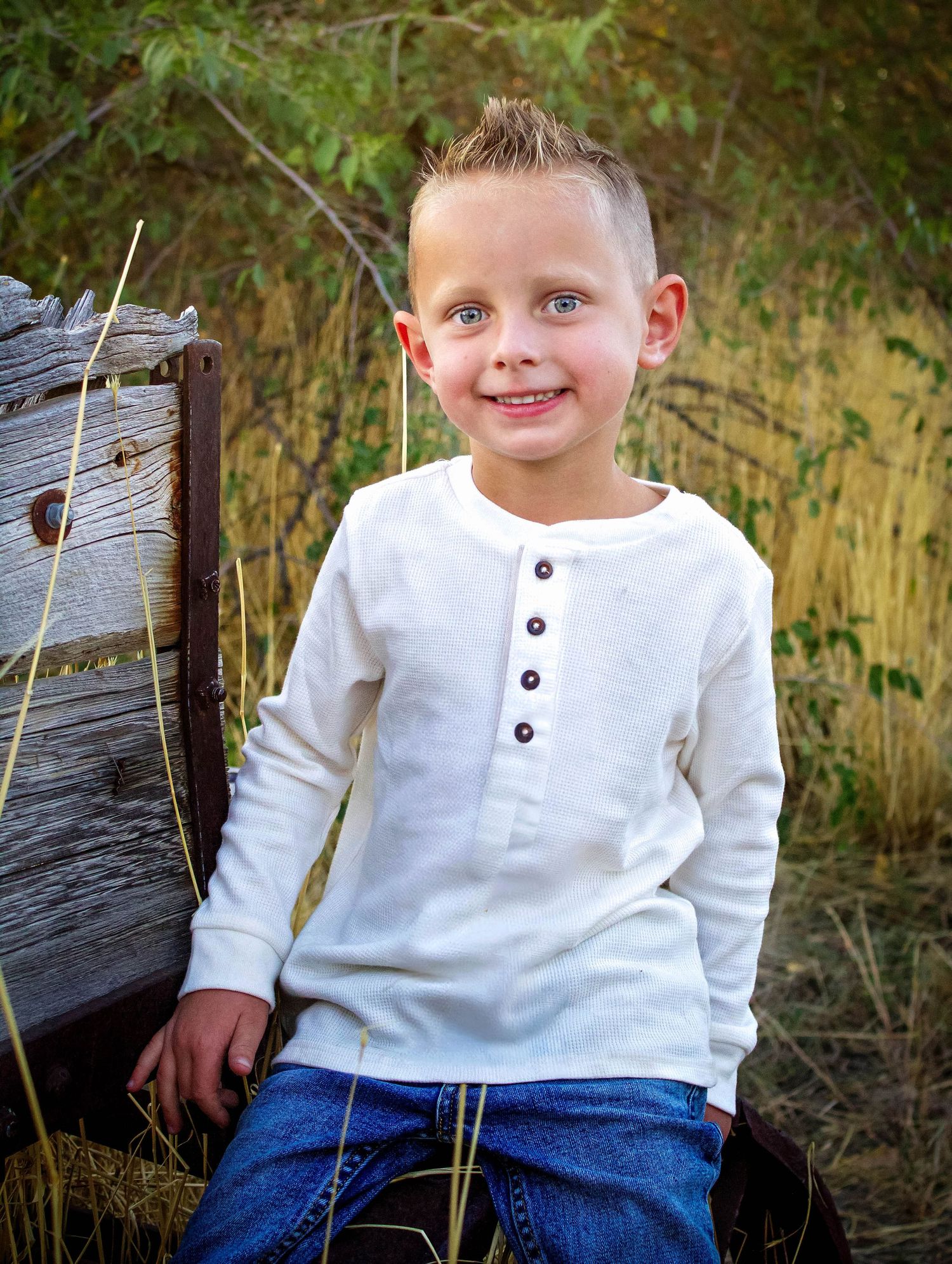 A child in white henley shirt and jeans sits on weathered wood in natural setting.
