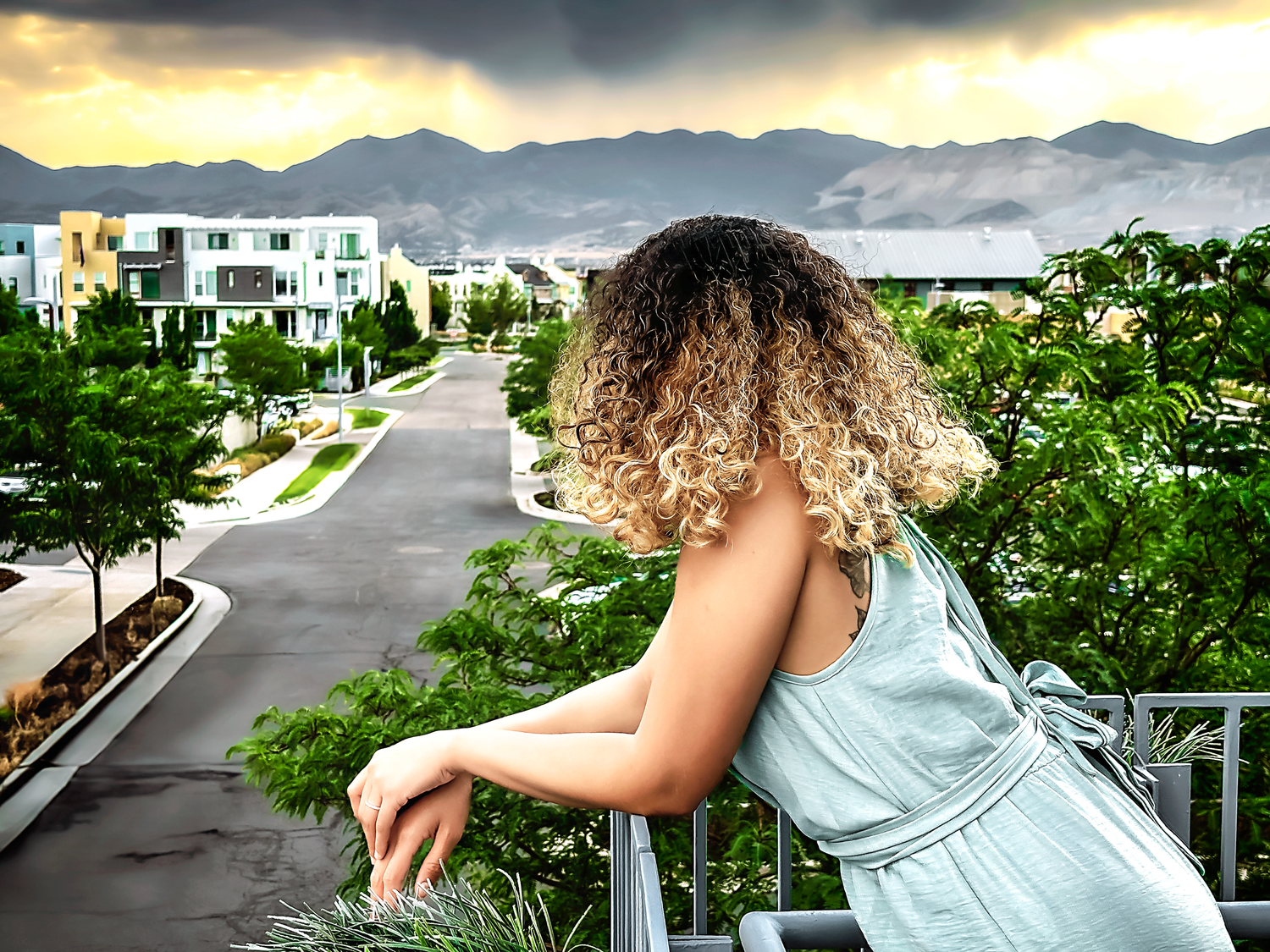 Person in blue dress sits on balcony railing with dramatic mountain sunset backdrop.