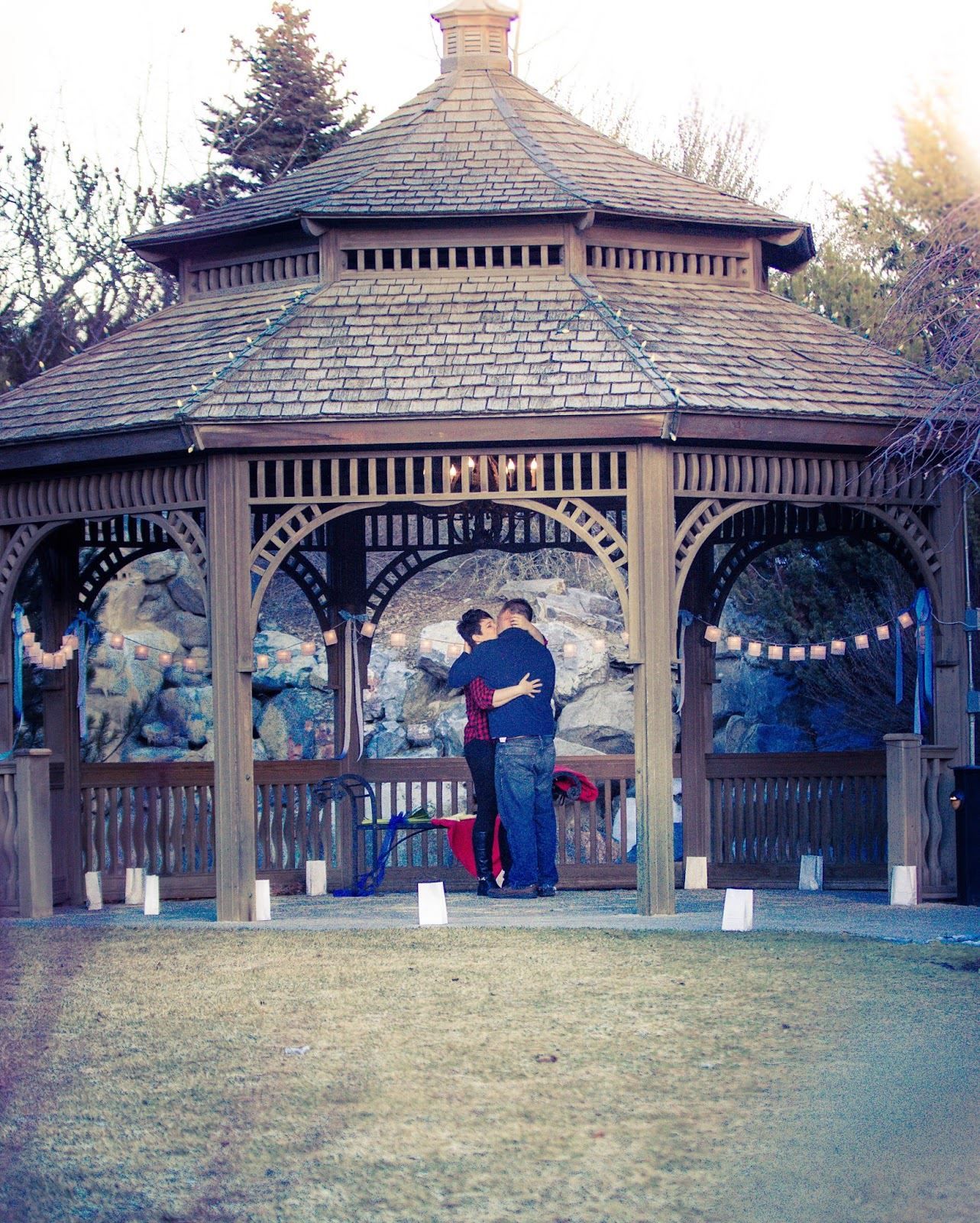 Wooden gazebo in park setting with ornate details and multiple connecting structures.