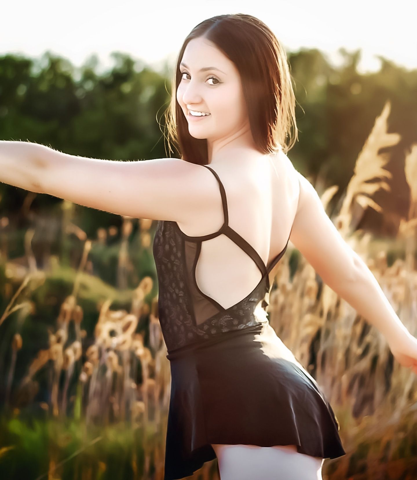 Woman in black dress poses gracefully in golden wheat field at sunset.