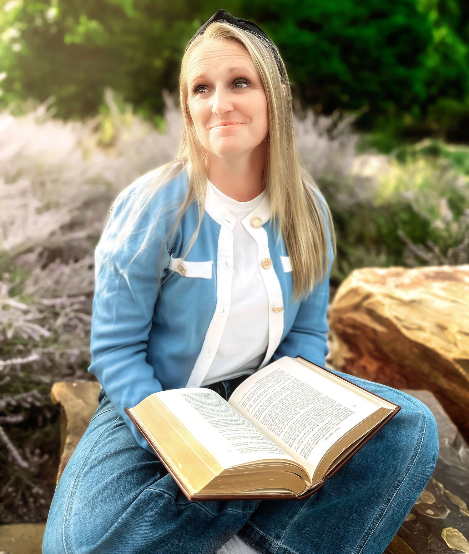 Person in blue sweater and jeans reads book in outdoor garden setting.