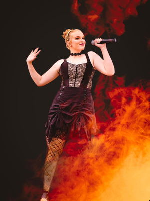 Theatrical performance shot with performer in corset dress against orange flame effects.