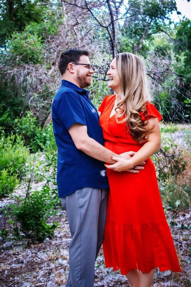 Couple in navy and red outfits embrace while laughing together during an outdoor photoshoot in a natural setting.