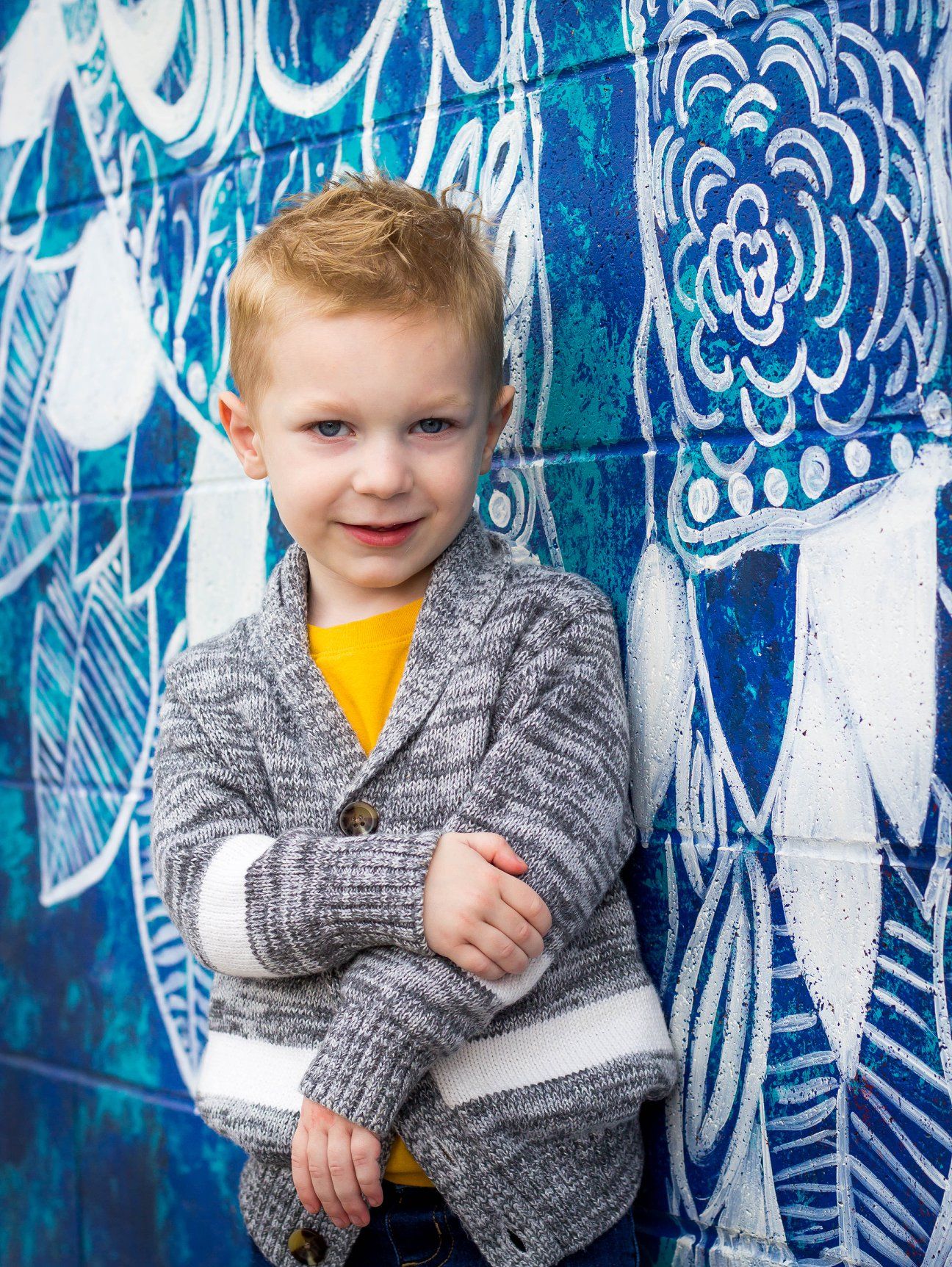 Child in gray cardigan and yellow shirt poses against blue artistic wall mural.