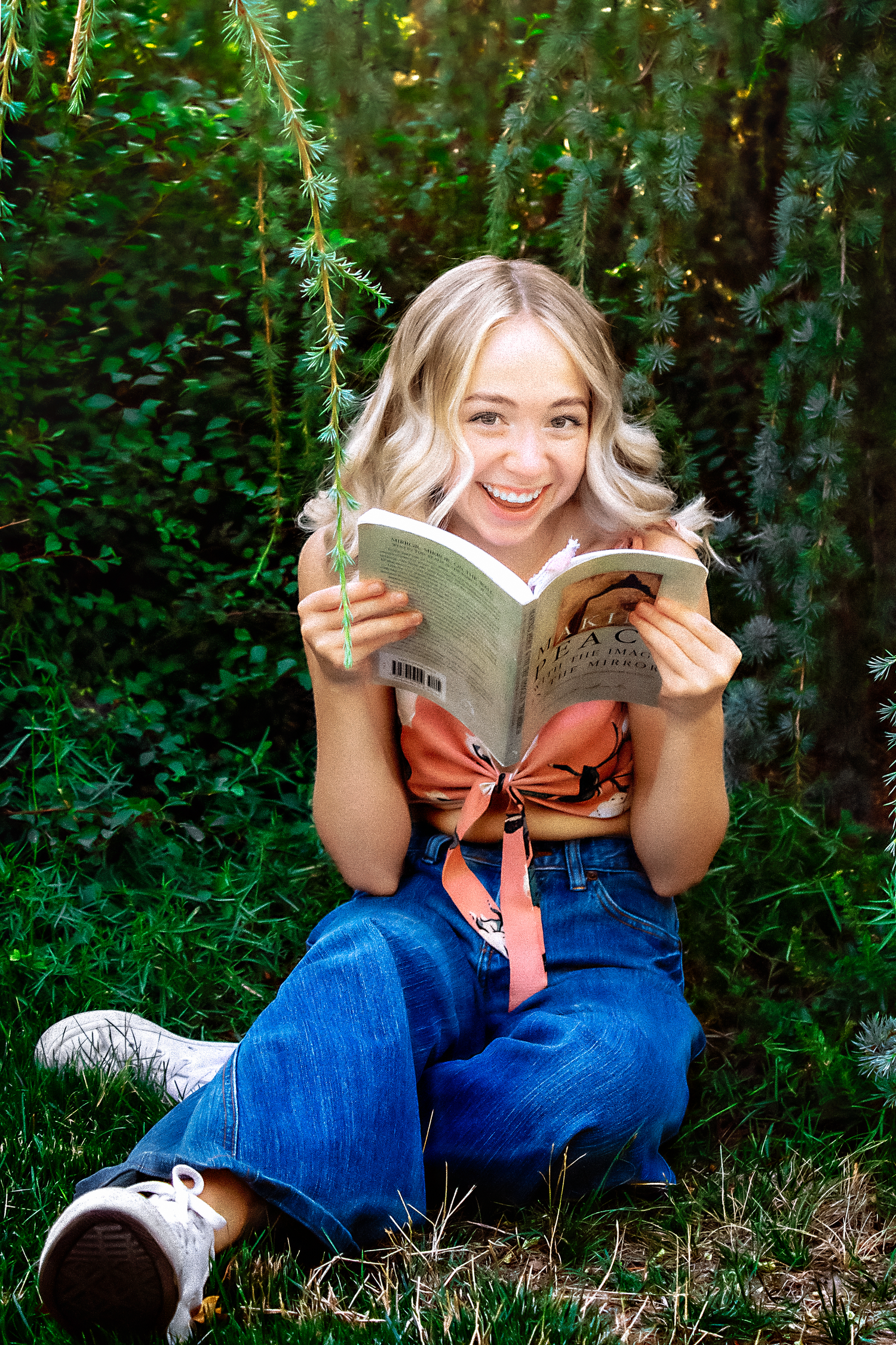 Reading a book while sitting on grass surrounded by greenery in a casual outdoor setting.