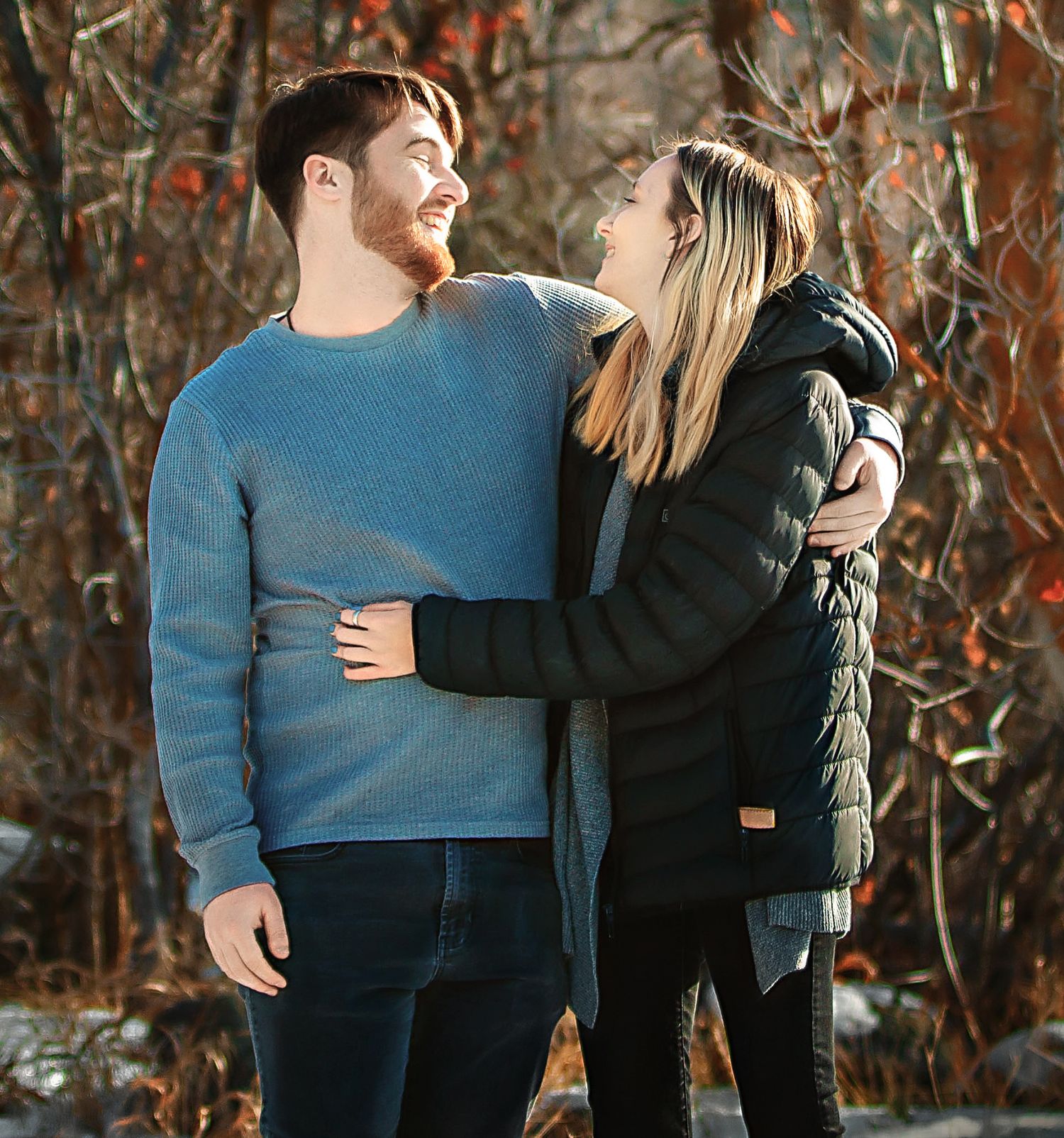 A couple embraces while standing in a forest with fall foliage in the background.