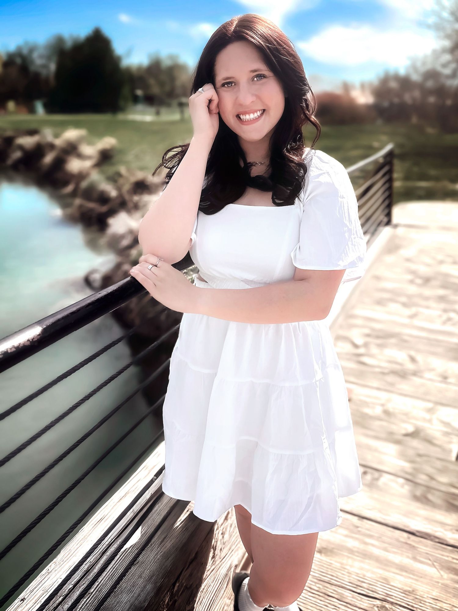 Person in white summer dress poses on wooden boardwalk near water.