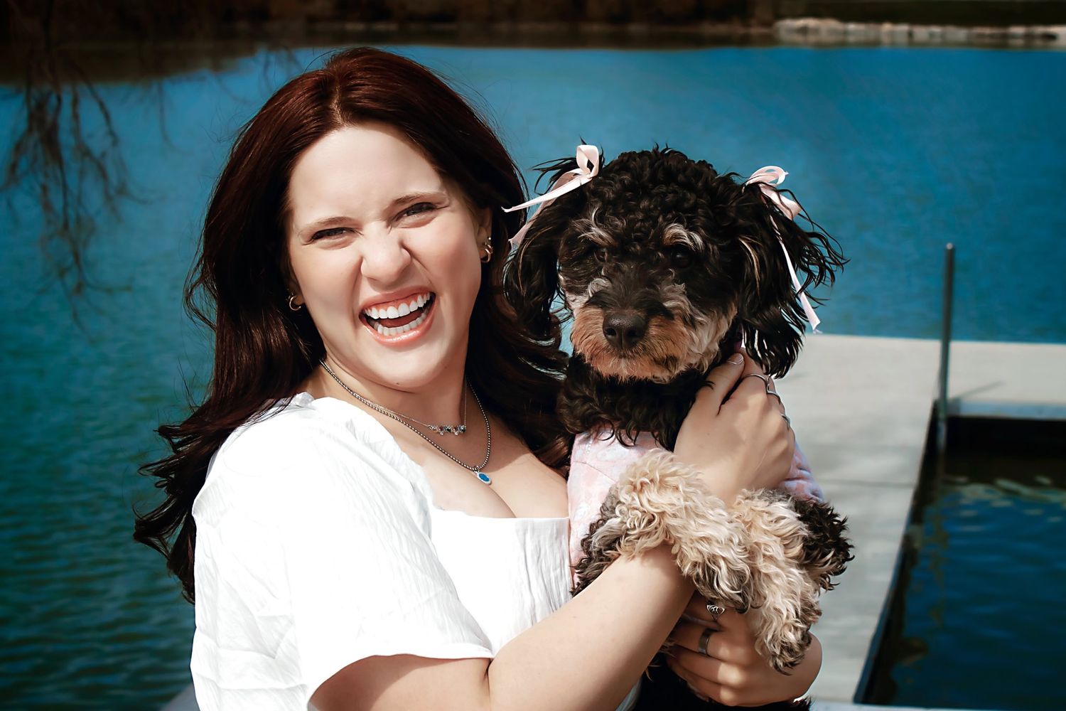 Person in white shirt holding small dog by lake during sunny day.