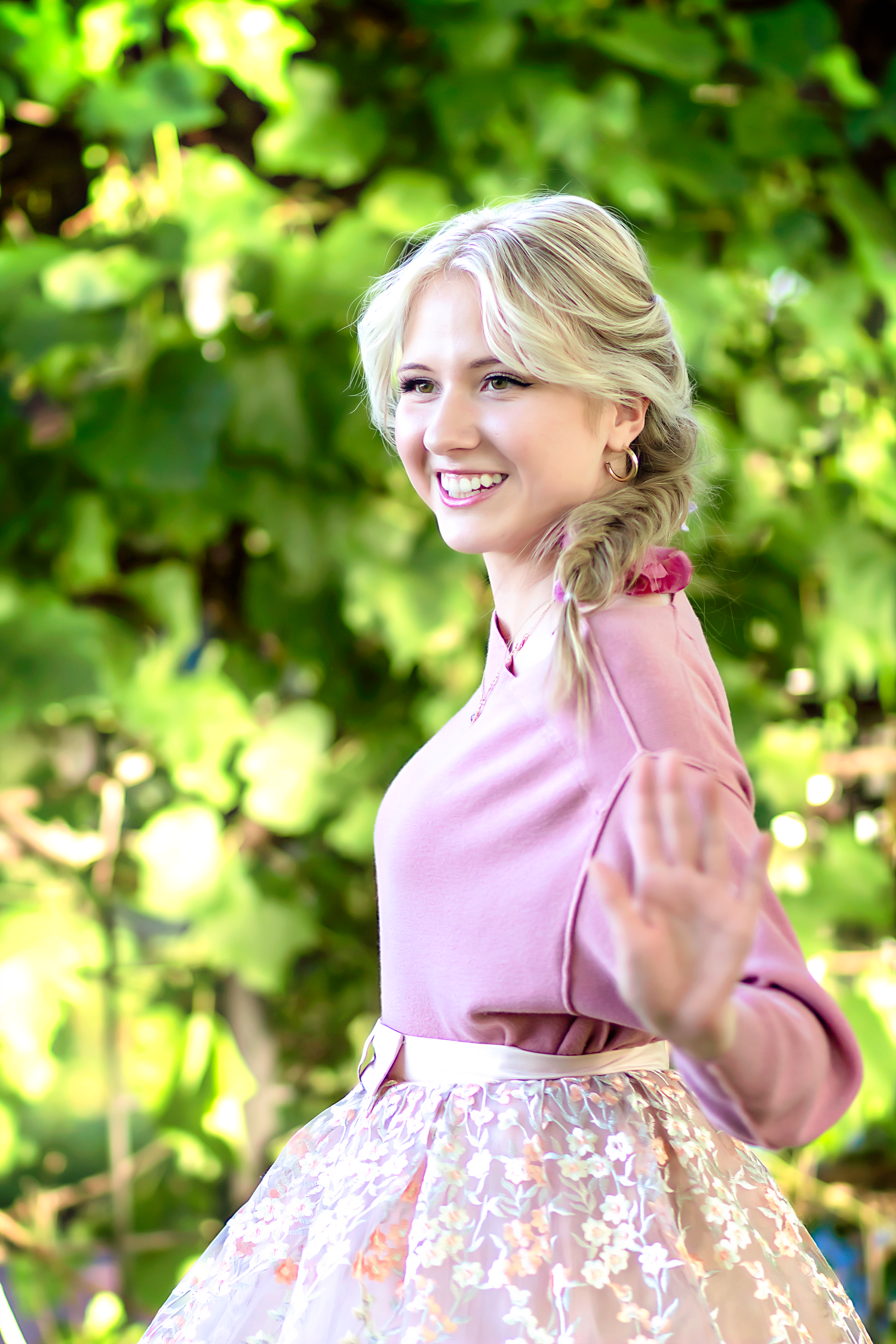 A dreamy outdoor portrait in a pink dress with floral details surrounded by sunlit green foliage.