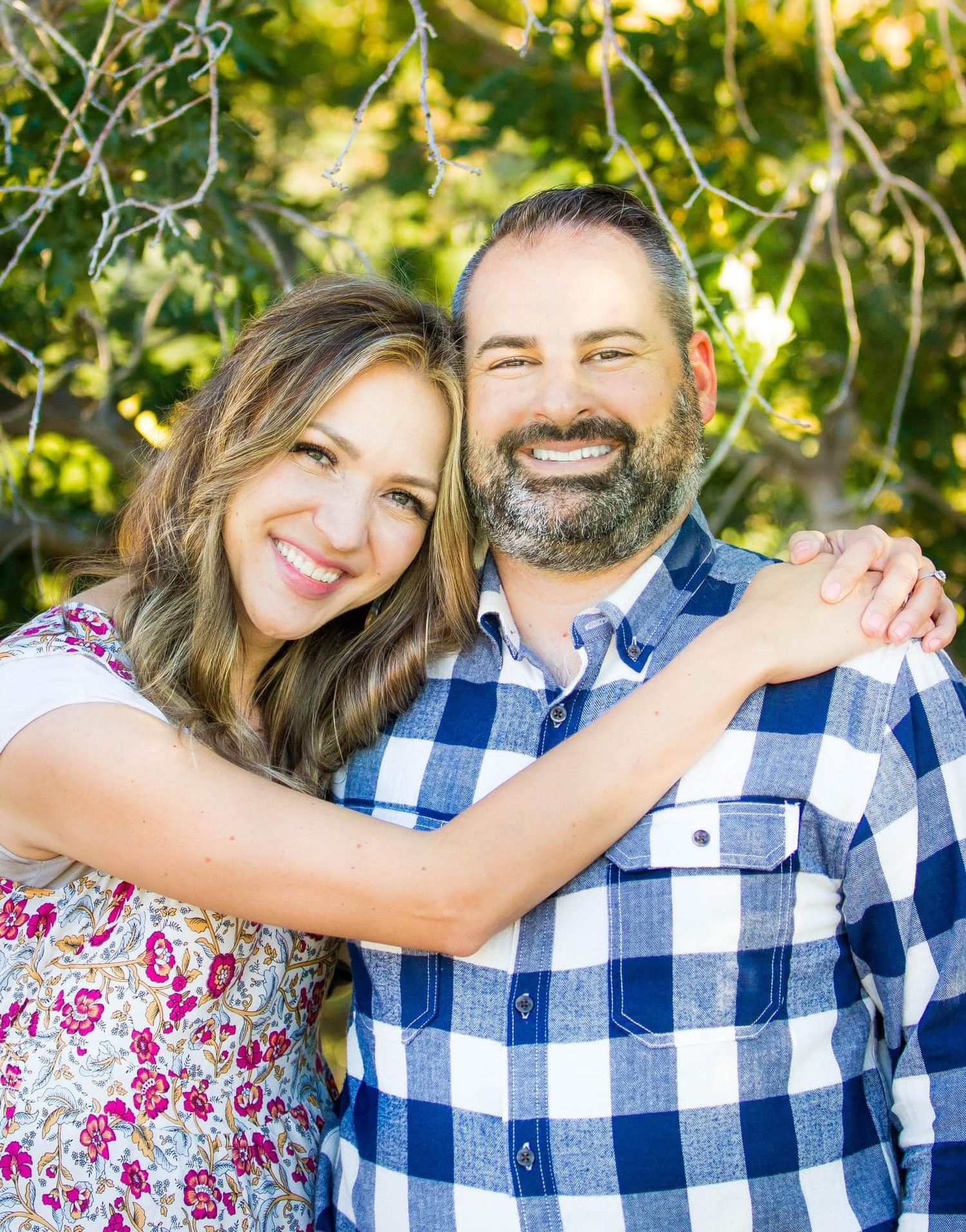 Couple in spring attire embracing outdoors with sunlit greenery in background.
