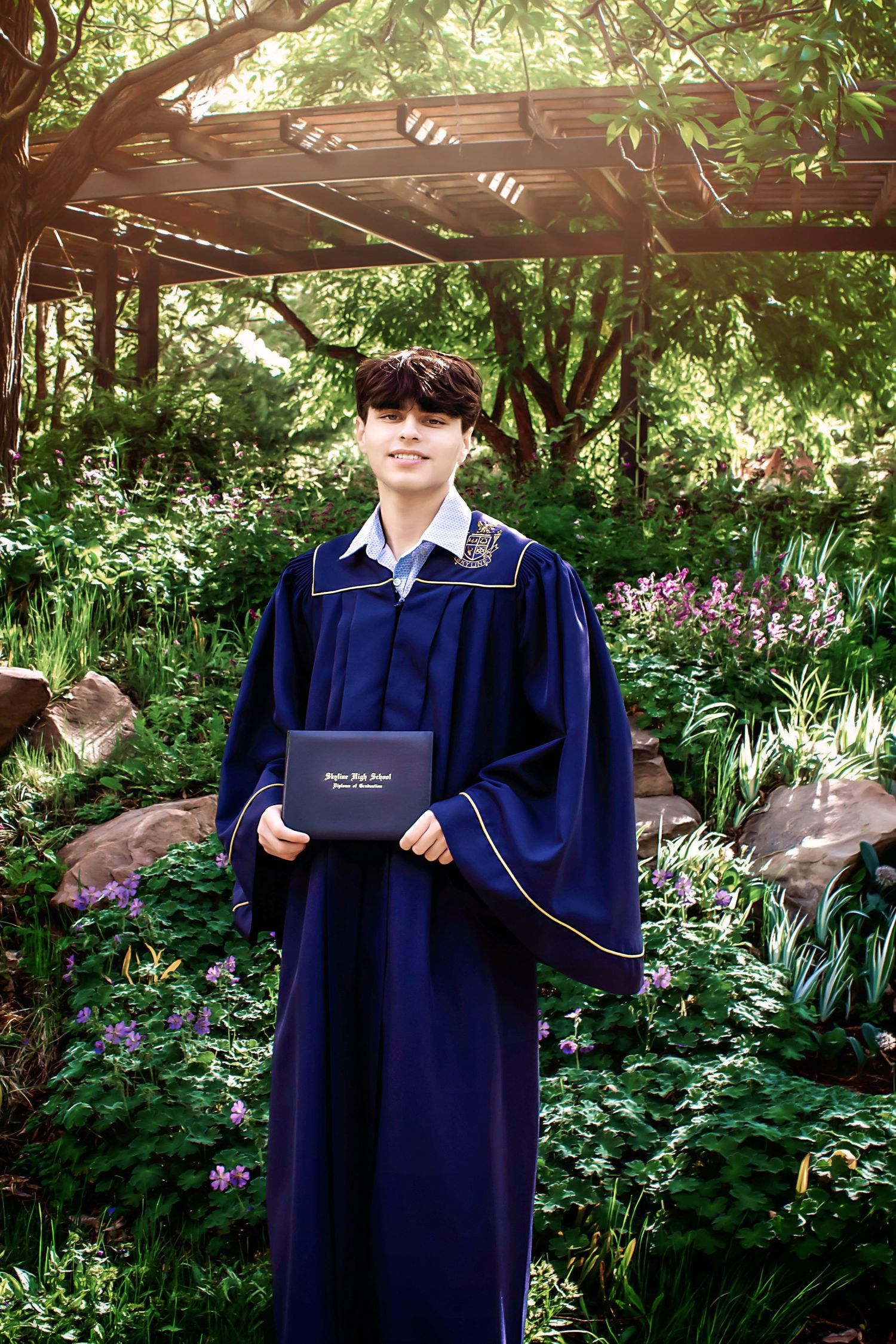 Graduate posing with diploma in garden setting under wooden pergola.