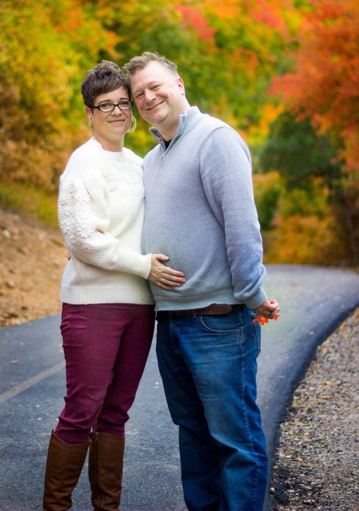 Couple in casual wear posing on path with vibrant fall colors in background.