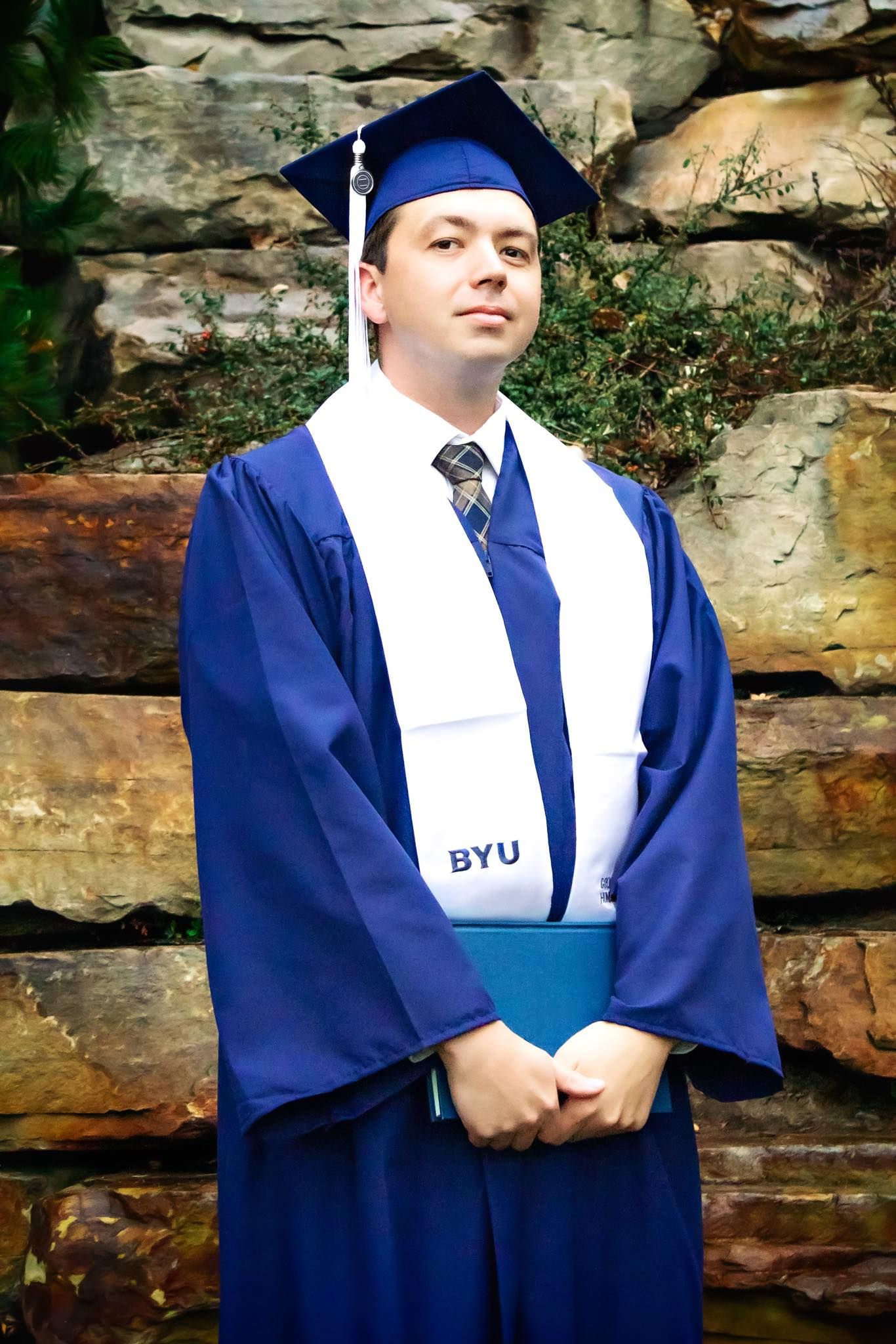 Graduate in blue BYU regalia stands against stone wall backdrop.