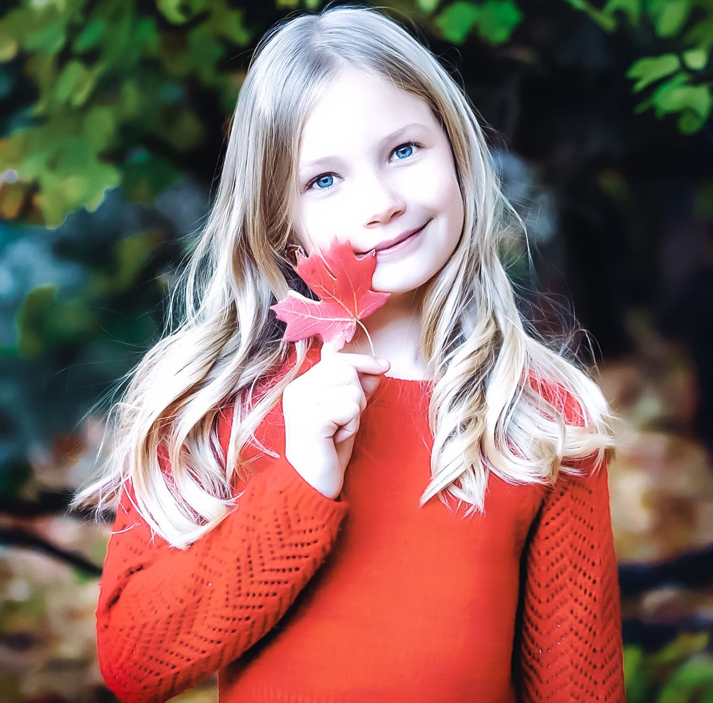 Young person in red sweater holding autumn maple leaf against fall foliage.