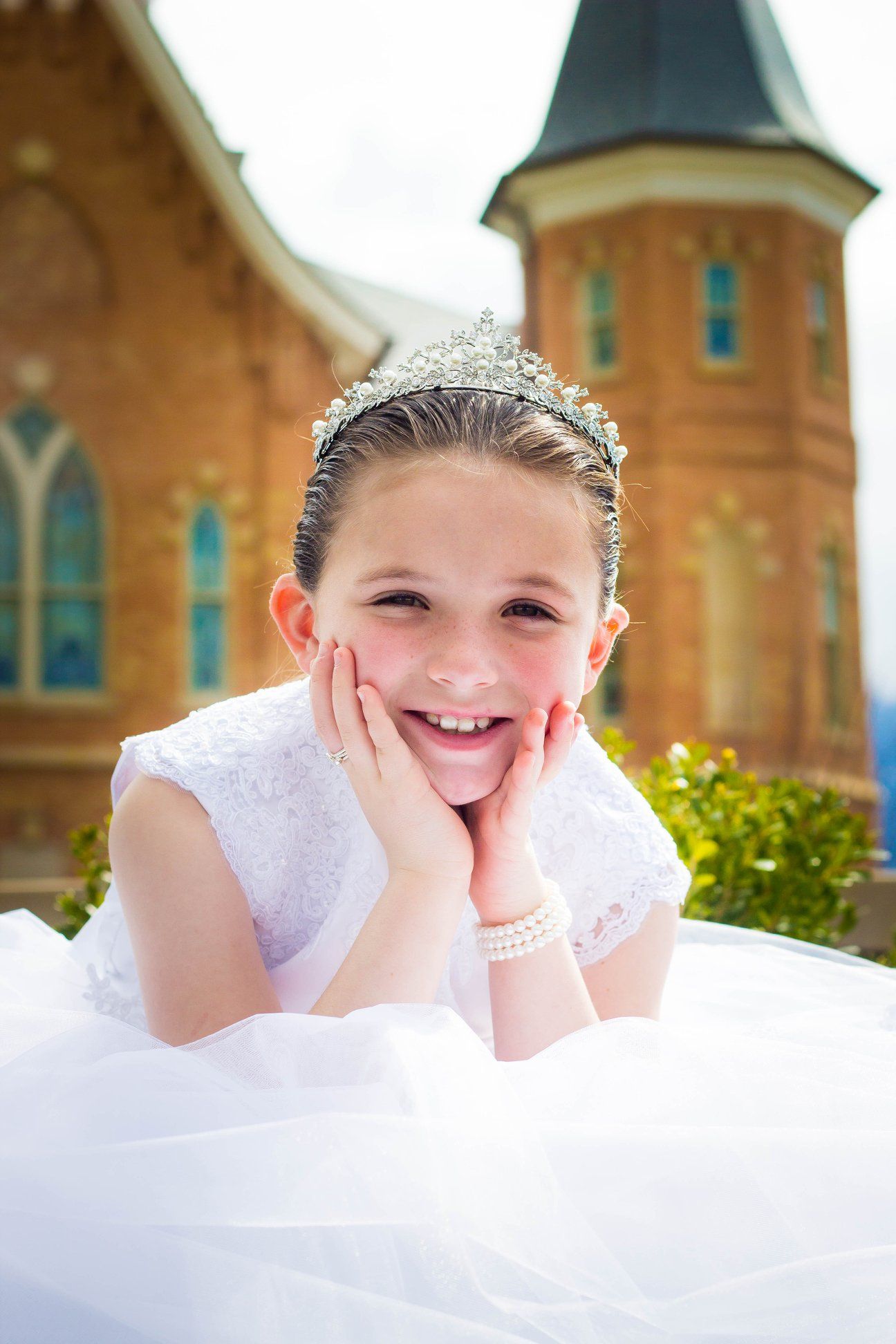 Person in white dress and tiara poses joyfully against brick church background.