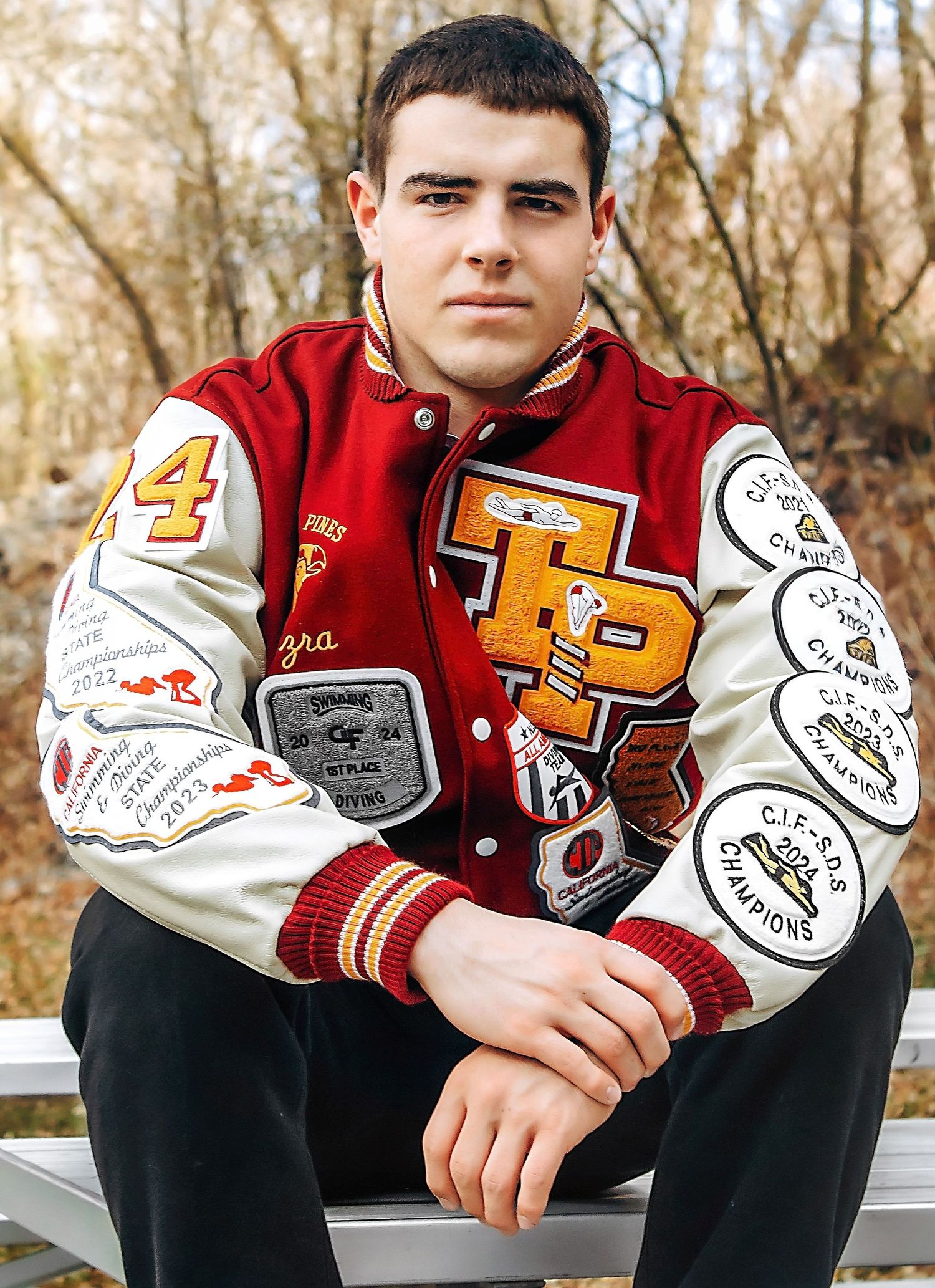 Person wearing varsity letterman jacket with patches sits outdoors in autumn setting.