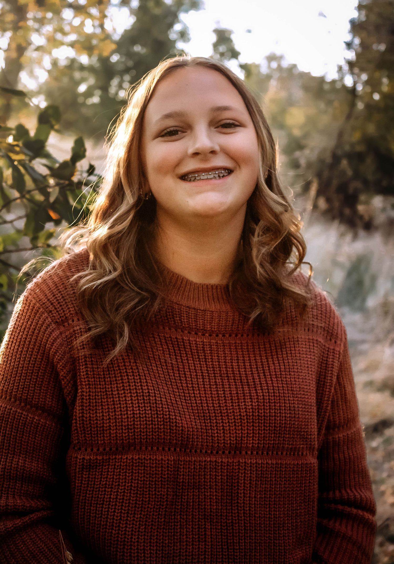 Person in brown knit sweater smiling outdoors with autumn foliage in background.