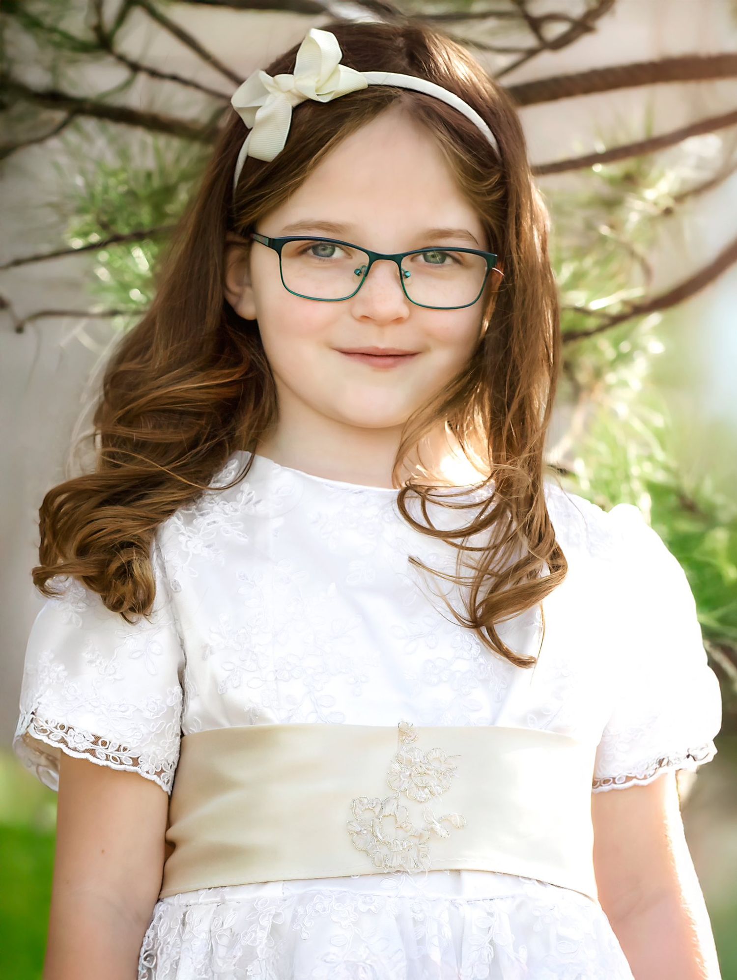 Young girl in white dress with bow headband and glasses poses for portrait against natural outdoor background.
