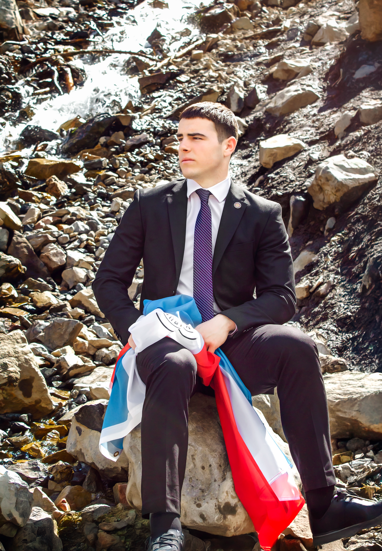 Person in business attire sitting thoughtfully on rocky terrain with flag nearby.