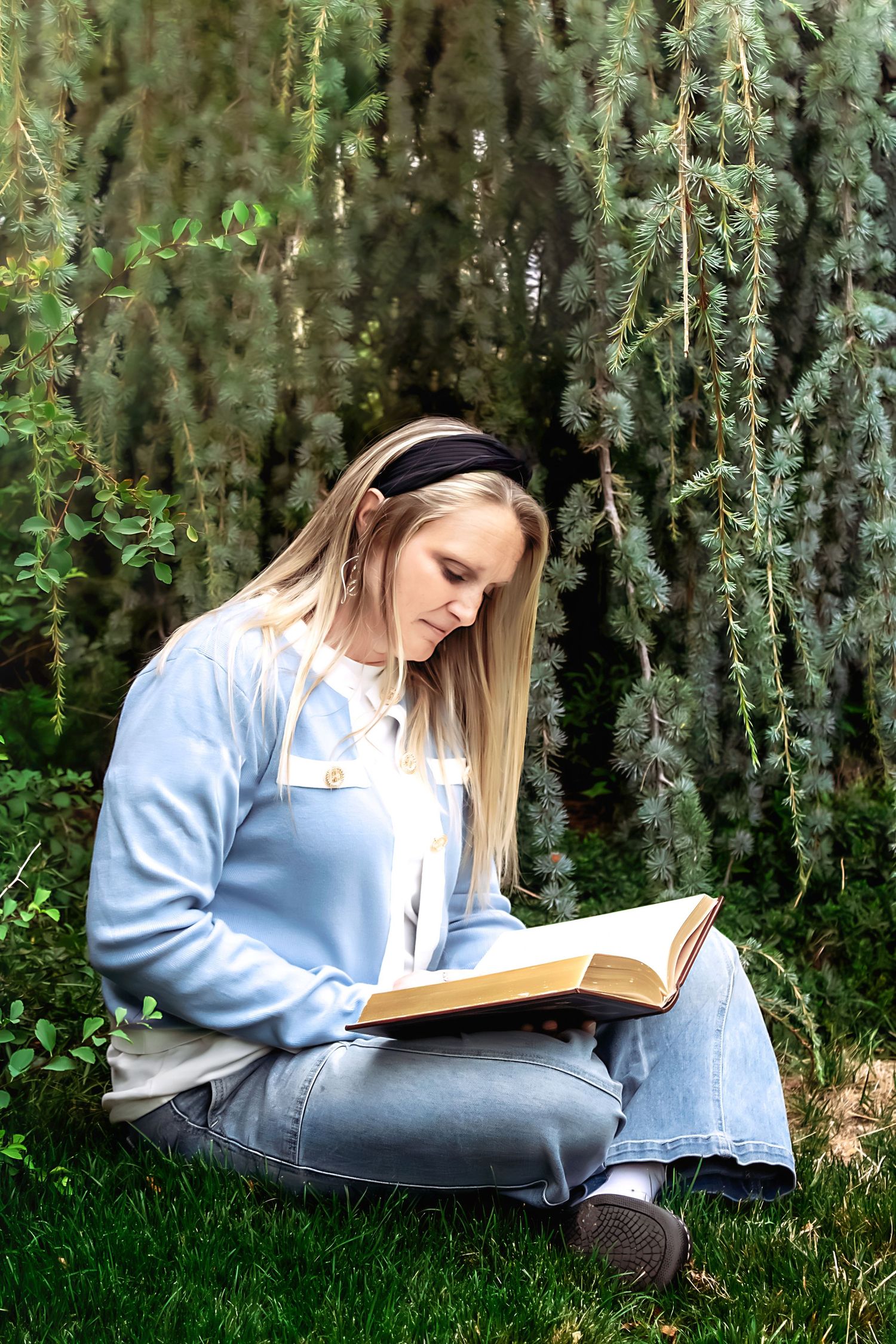 A person in a blue sweater reads a book while sitting on grass in a garden setting.