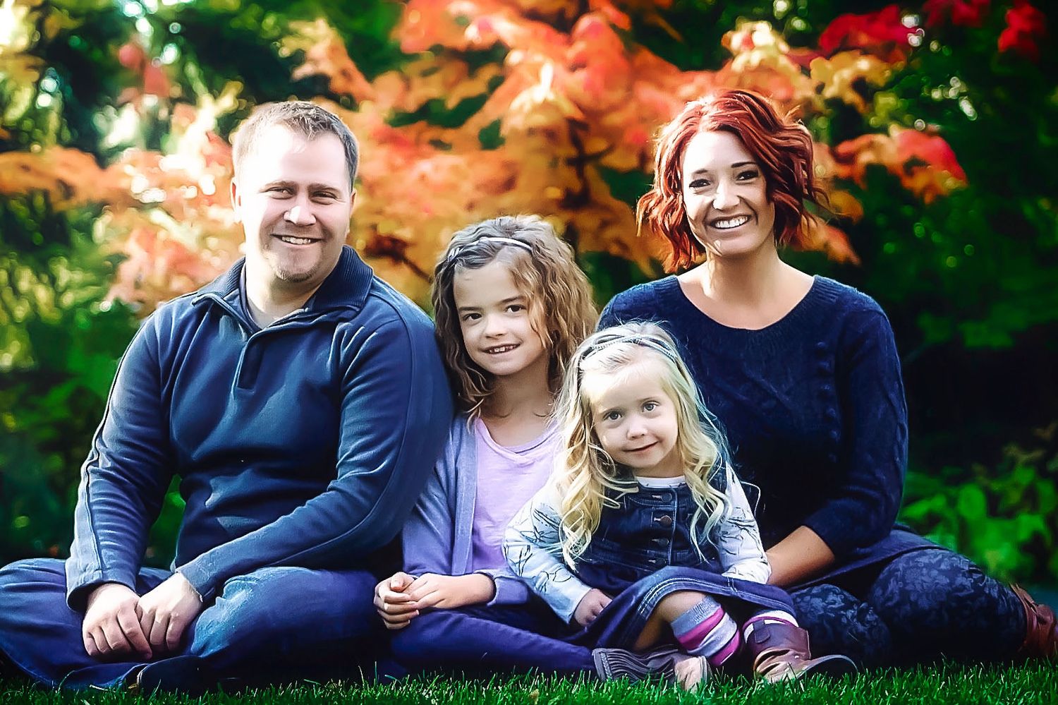 A family of four sits together on grass with colorful autumn trees in the background.